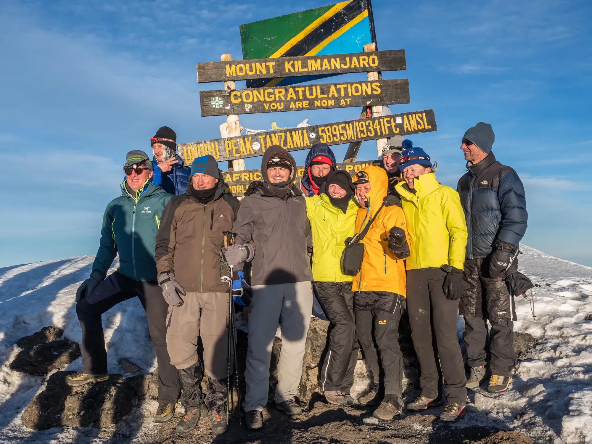 Eine Hauser-Trekkinggruppe posiert glücklich am Gipfelschild des Uhuru Peak auf dem Kilimanjaro in Tansania.
