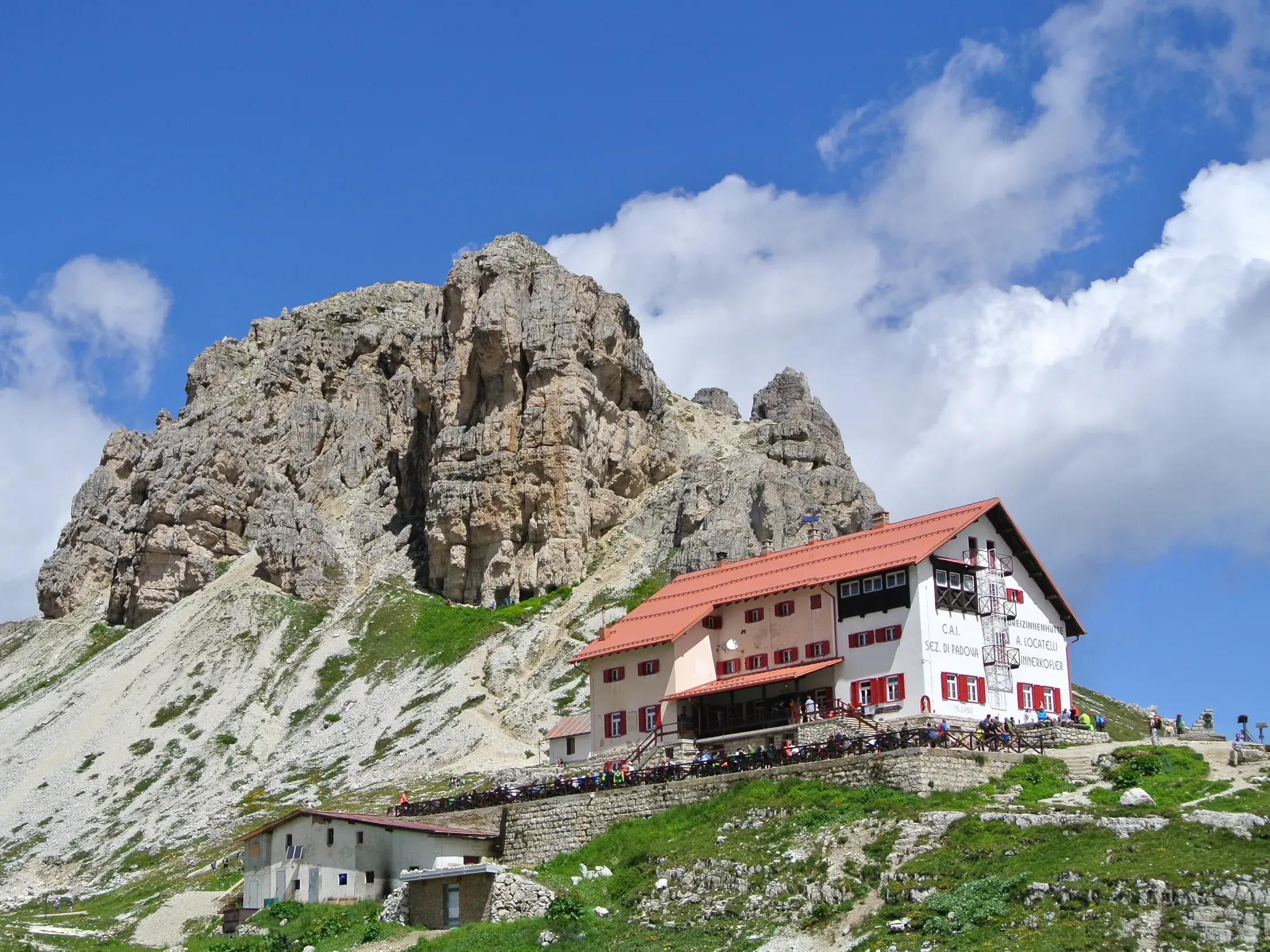 Die Drei Zinnen Hütte in den Sextner Dolomiten, wo Wanderer vor einer imposanten Felskulisse eine sonnige Pause einlegen.
