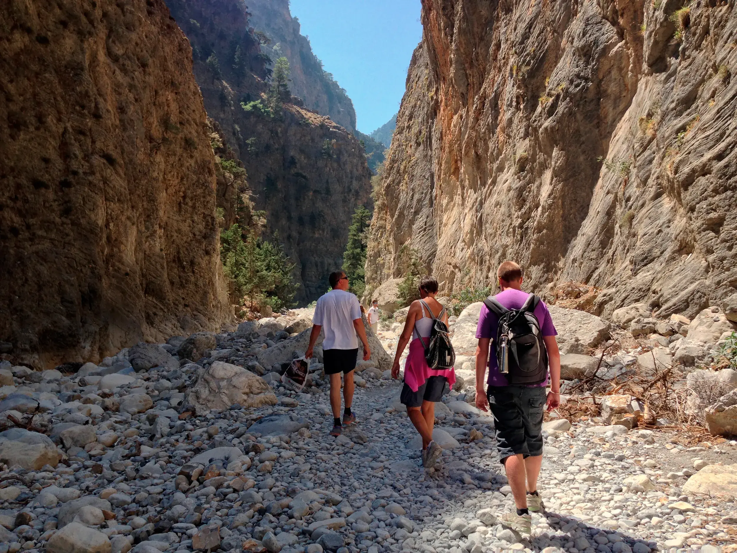Drei Wanderer einer Reisegruppe durchqueren auf einem steinigen Pfad die imposante Samariaschlucht auf Kreta, umgeben von hohen Felswänden.