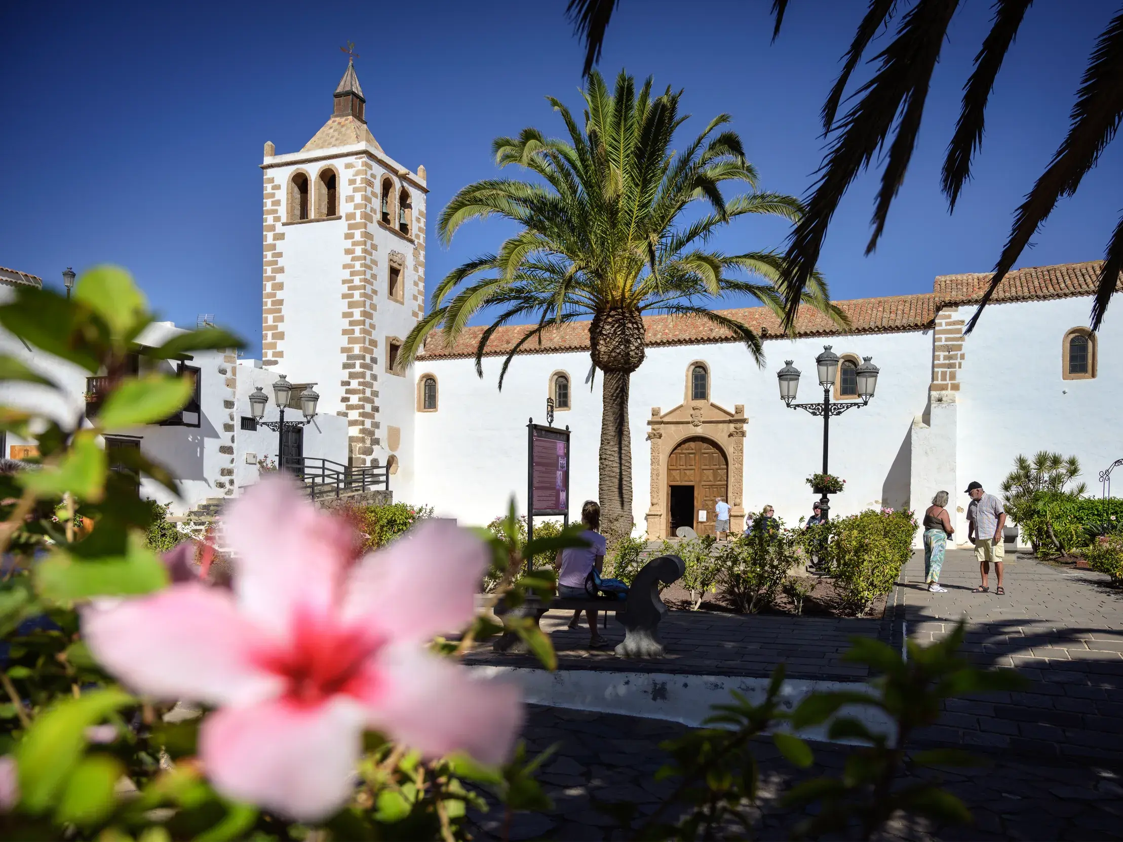 Die historische Kirche Santa María in Betancuria auf Fuerteventura, umgeben von Palmen, mit Besuchern auf dem sonnigen Kirchplatz.