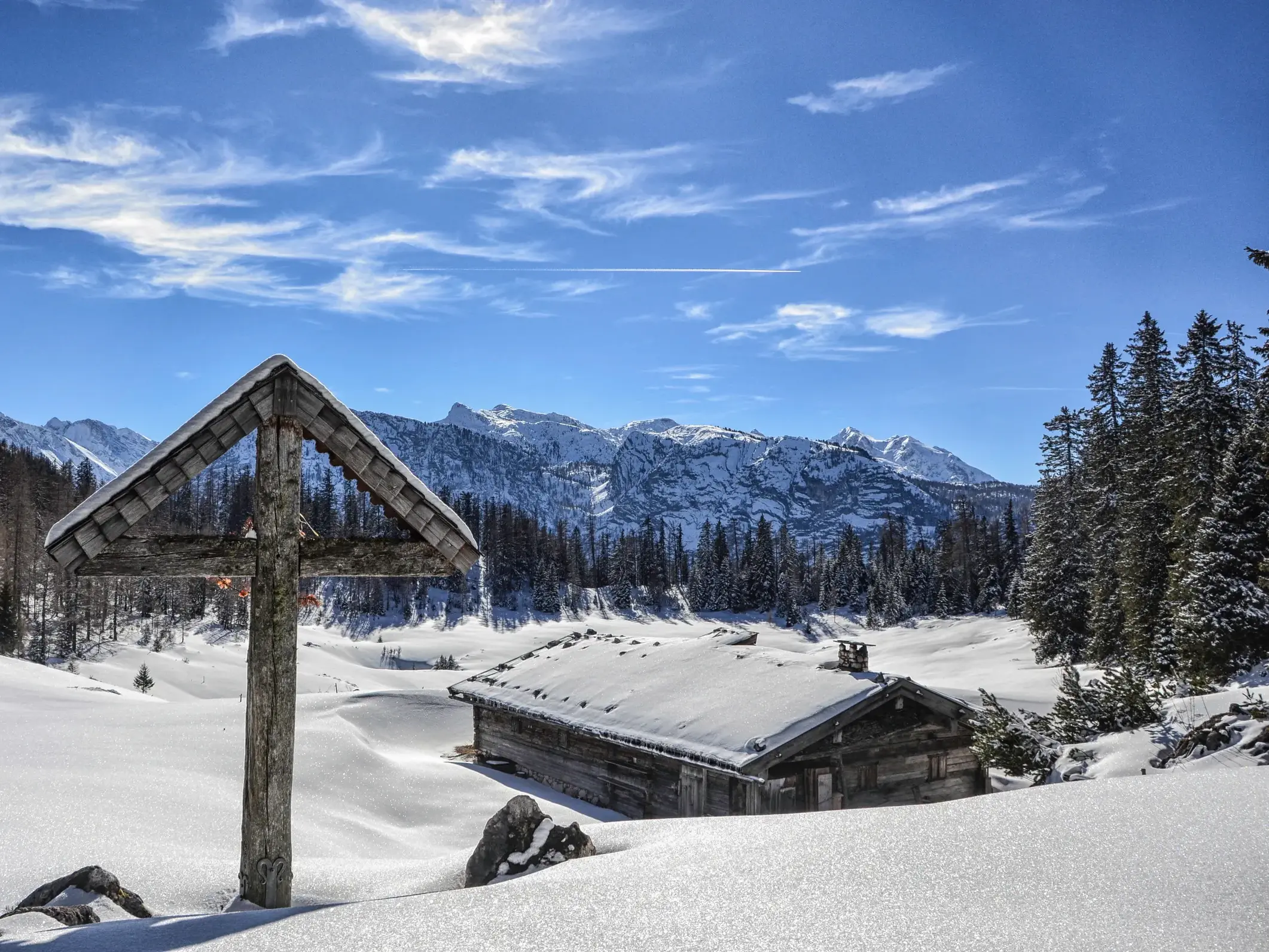 Wegkreuz mit Holzdach und verschneite Almhütte vor dem Panorama der Berchtesgadener Alpen an einem sonnigen Wintertag.