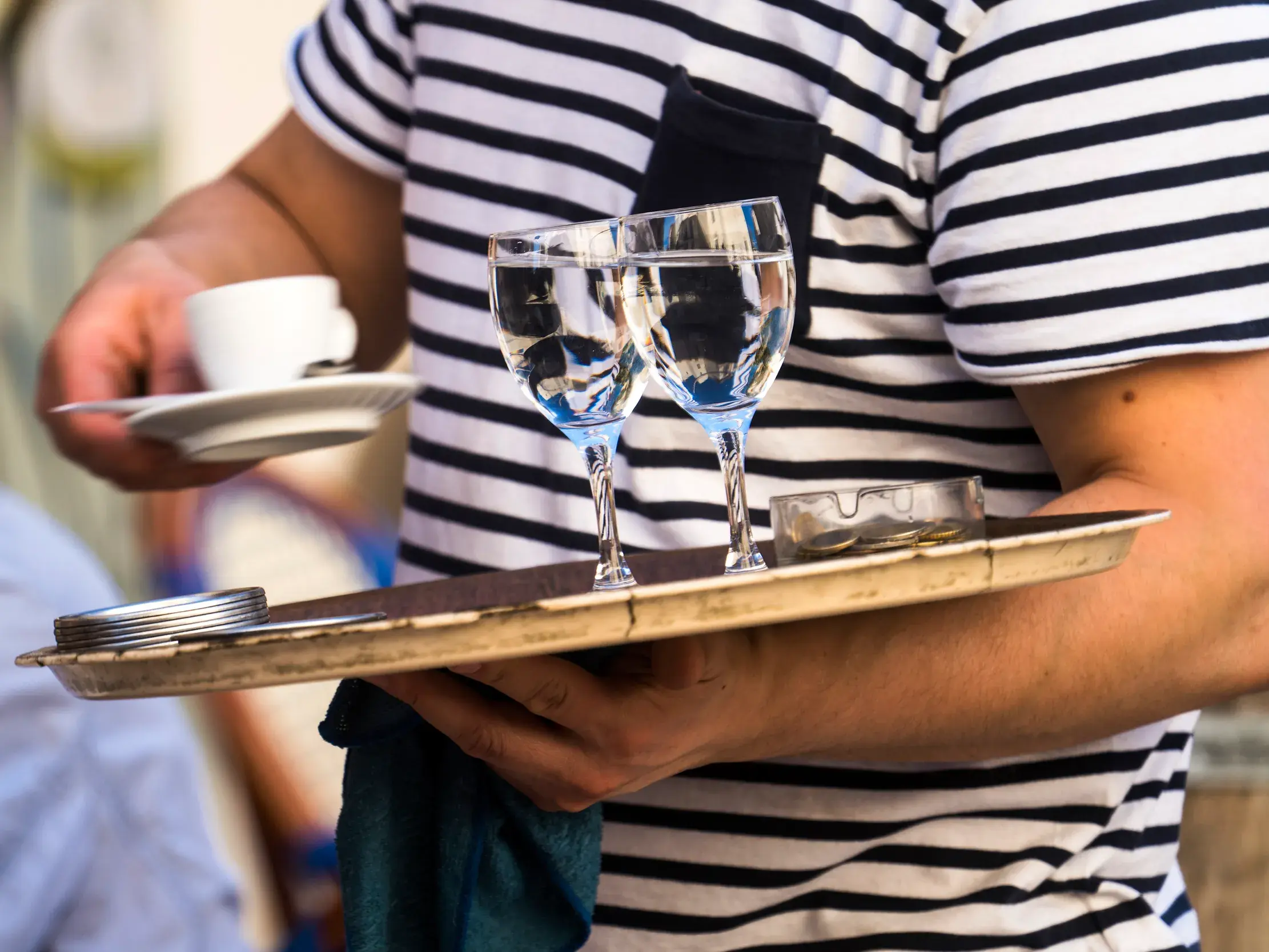 Ein Kellner in einem typisch französischen Streifenshirt trägt ein Tablett mit Wassergläsern und eine Tasse Kaffee.