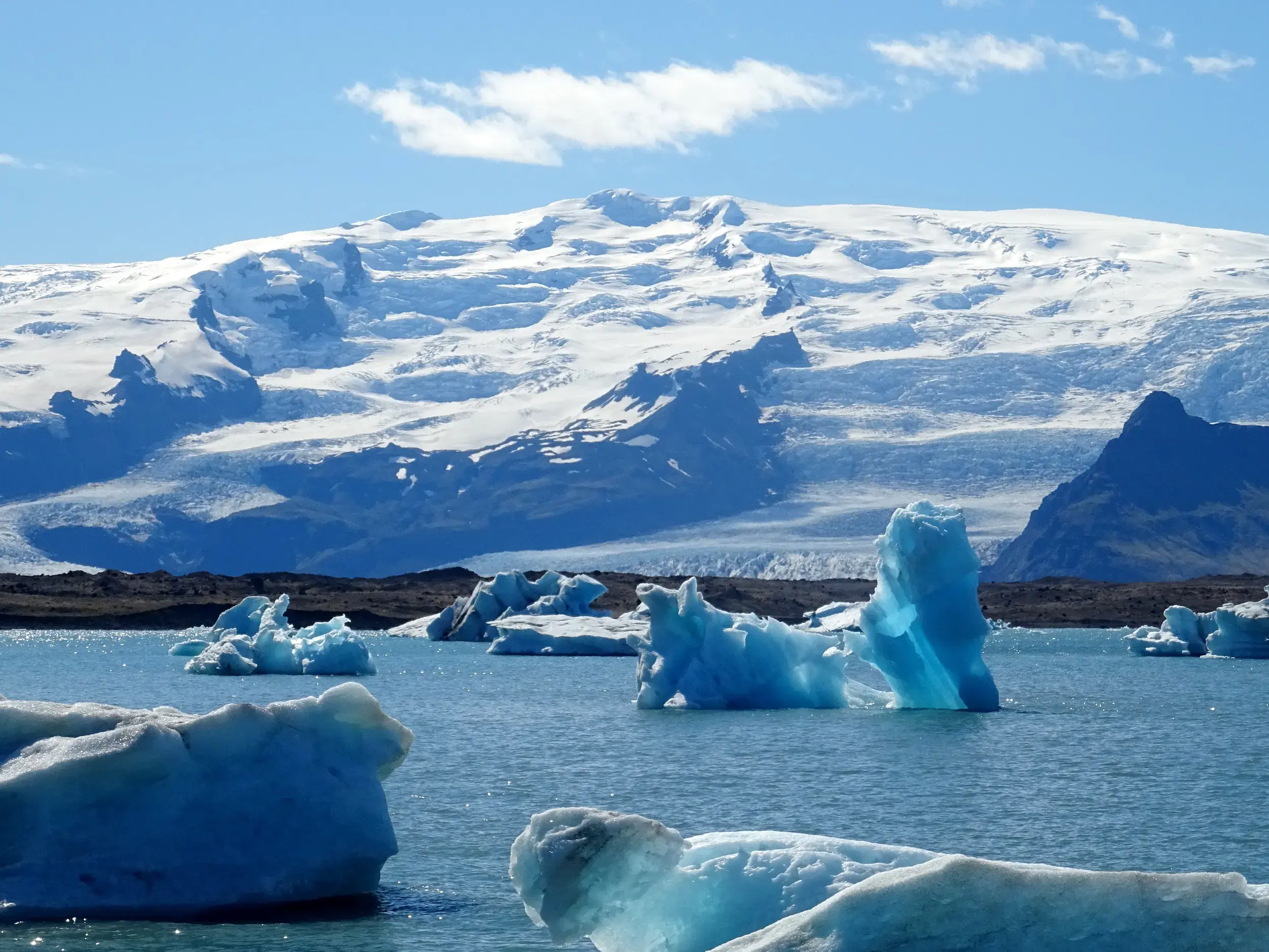 Blau schimmernde Eisberge treiben in der Gletscherlagune Jökulsárlón vor der Eiskappe des Vatnajökull an einem sonnigen Tag.