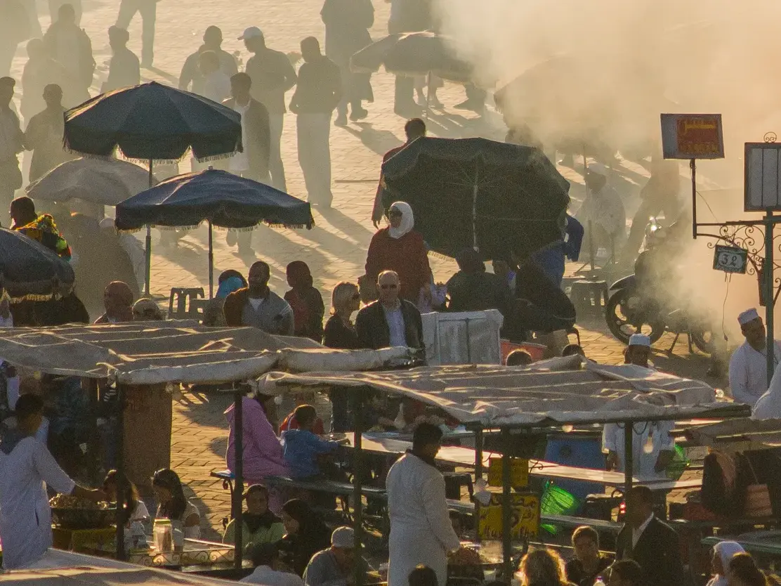 Zahlreiche Menschen an den dampfenden Garküchen auf dem Djemaa el Fna in Marrakesch in der goldenen Abendsonne.