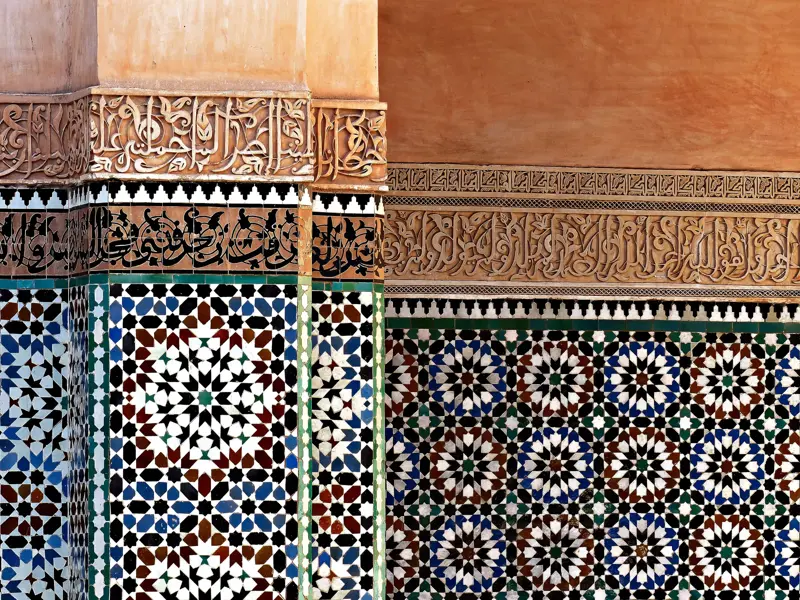 Nahaufnahme der filigranen Mosaikkunst und Stuckverzierungen mit Kalligrafie an der Wand der Medersa Ben Youssef in Marrakesch.