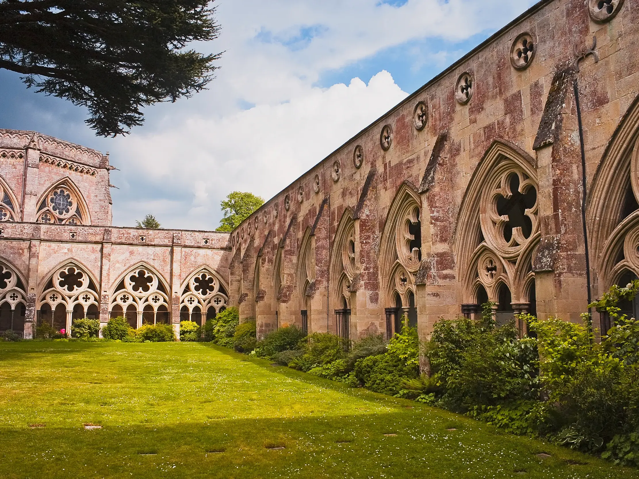 Gotischer Kreuzgang der Kathedrale von Salisbury in Südengland, dessen Arkaden einen grünen, sonnigen Innenhof umschließen.