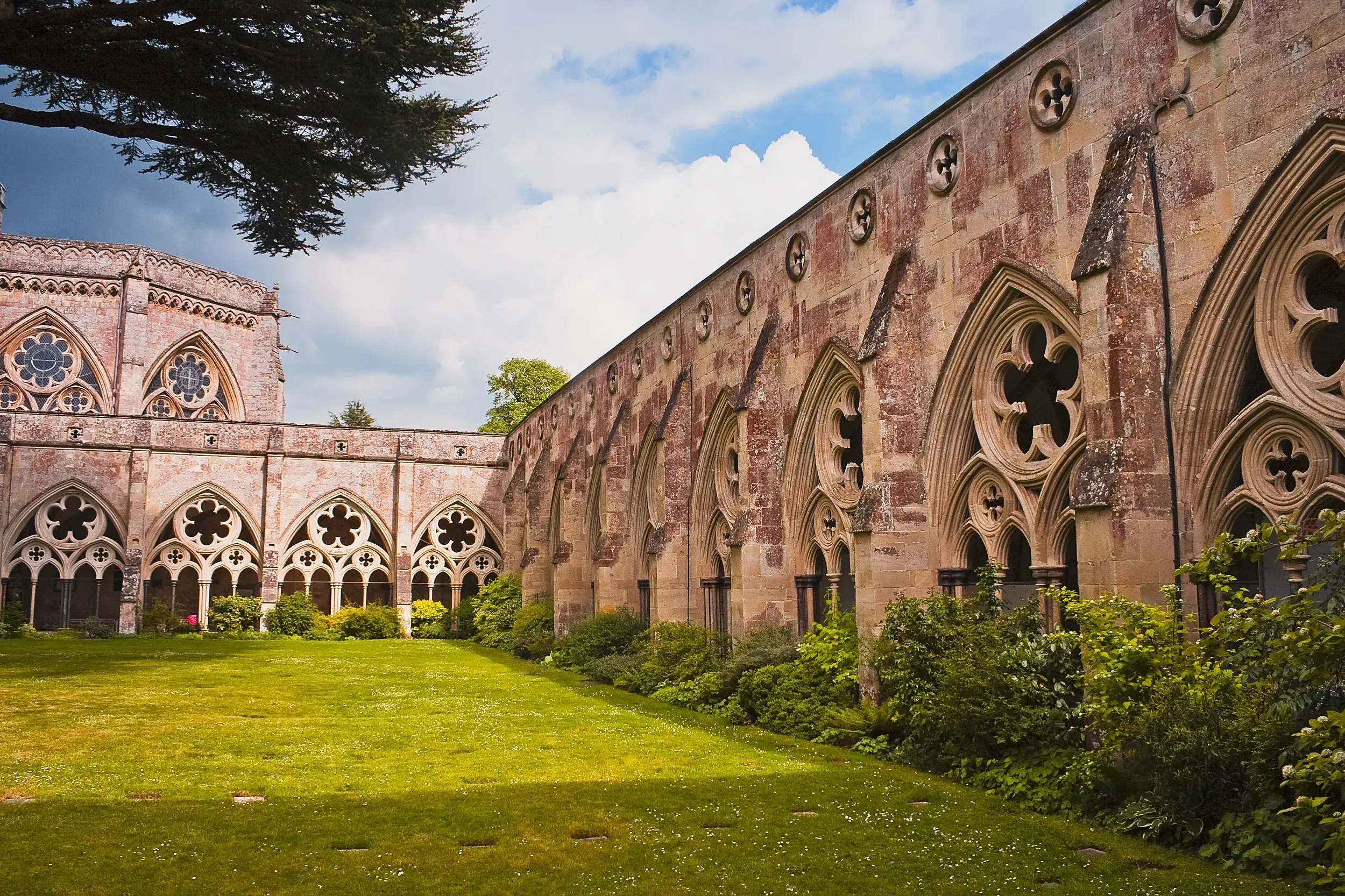 Gotischer Kreuzgang der Kathedrale von Salisbury in Südengland, dessen Arkaden einen grünen, sonnigen Innenhof umschließen.