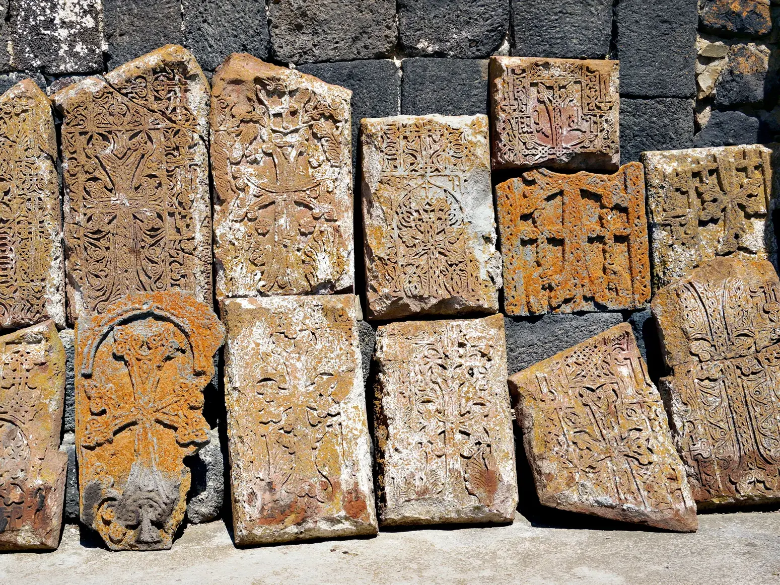 Zahlreiche historische Chatschkars, armenische Kreuzsteine mit filigranen Gravuren, lehnen an einer dunklen Steinmauer bei einem Kloster in Armenien.