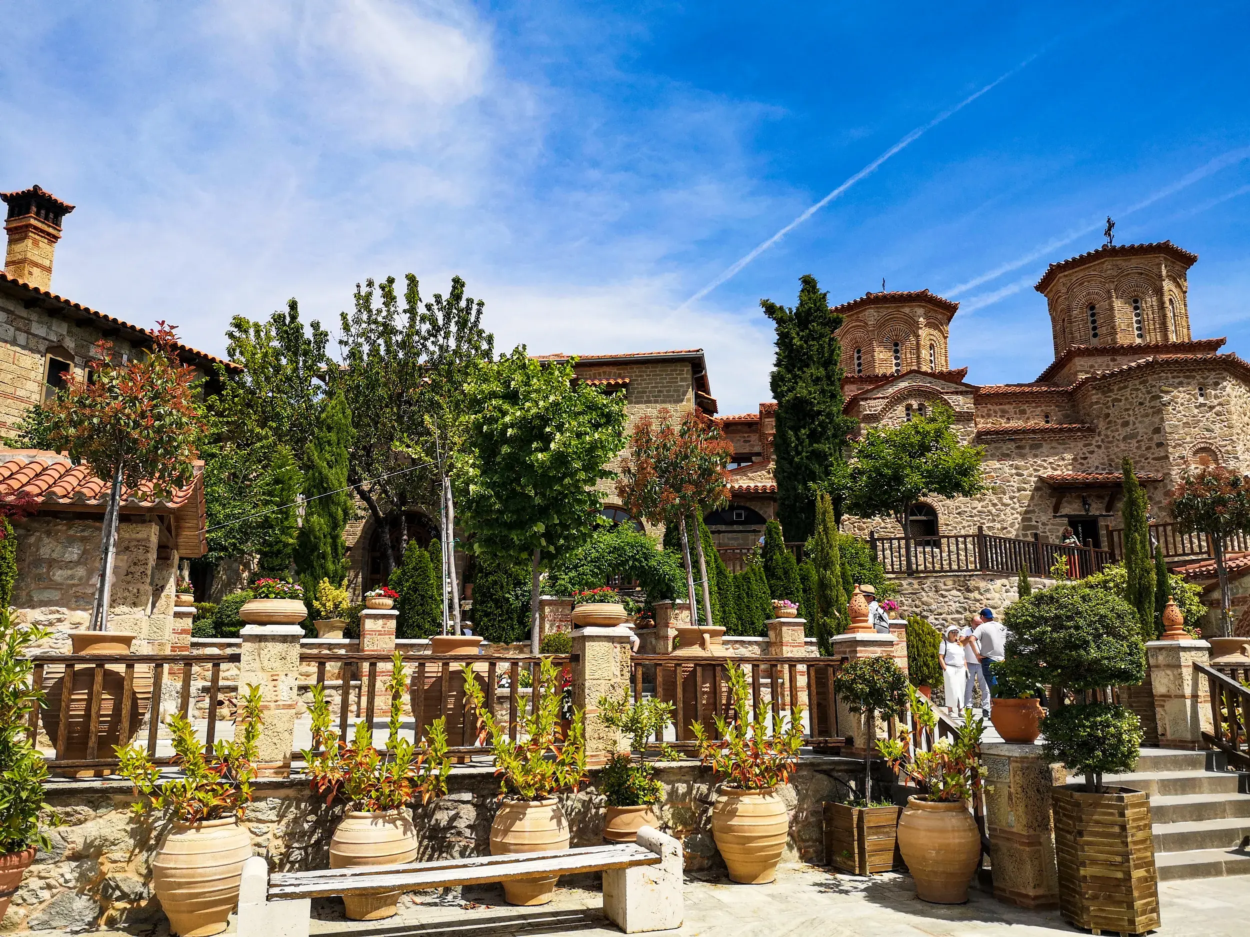 Sonniger Innenhof des Metamorphosis Klosters bei Meteora, umgeben von Steinmauern, grünen Pflanzen und byzantinischen Türmen.
