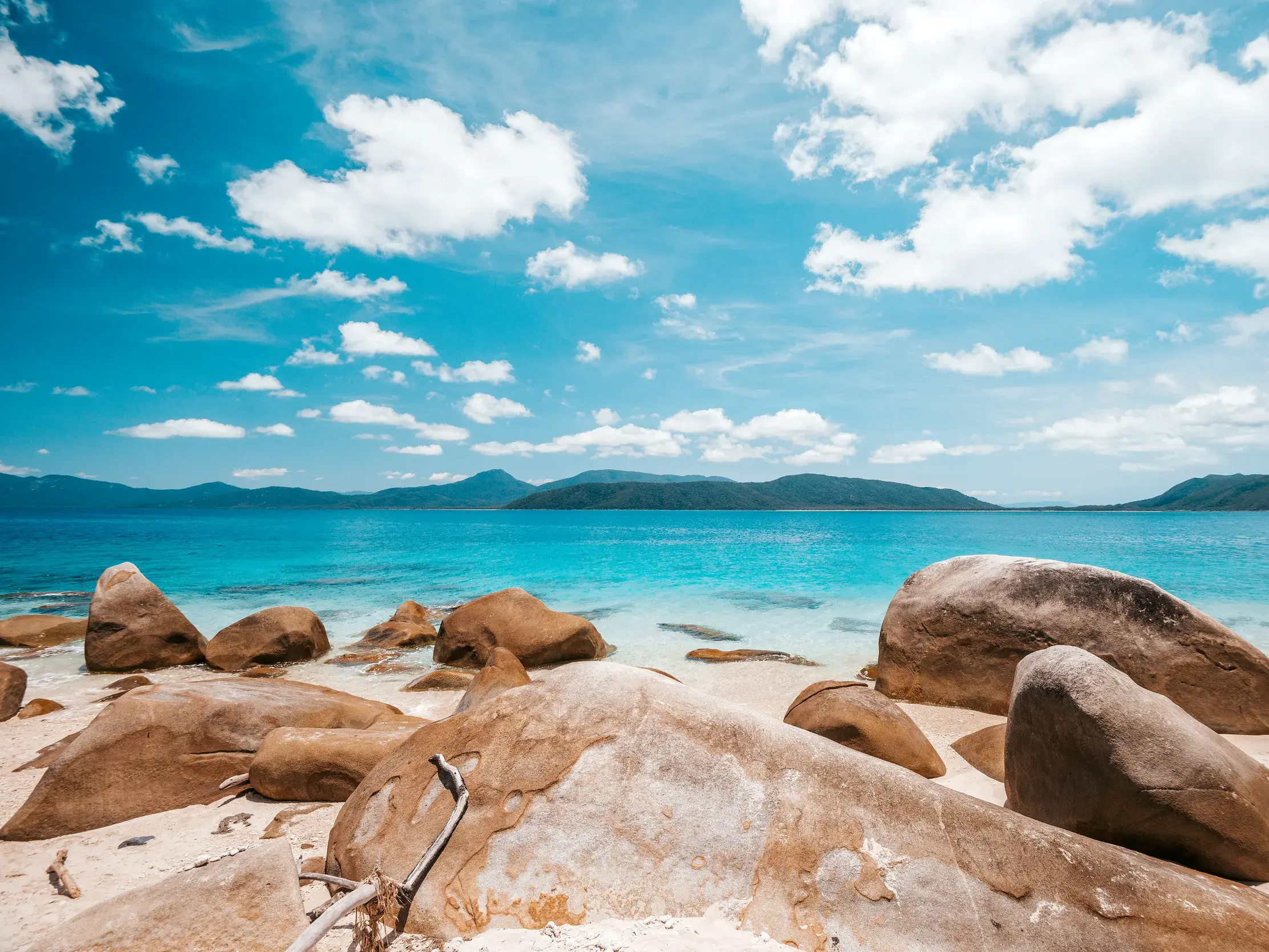 Große Granitfelsen am Sandstrand einer Bucht in Queensland mit türkisem Wasser und grünen Inseln unter blauem Himmel.