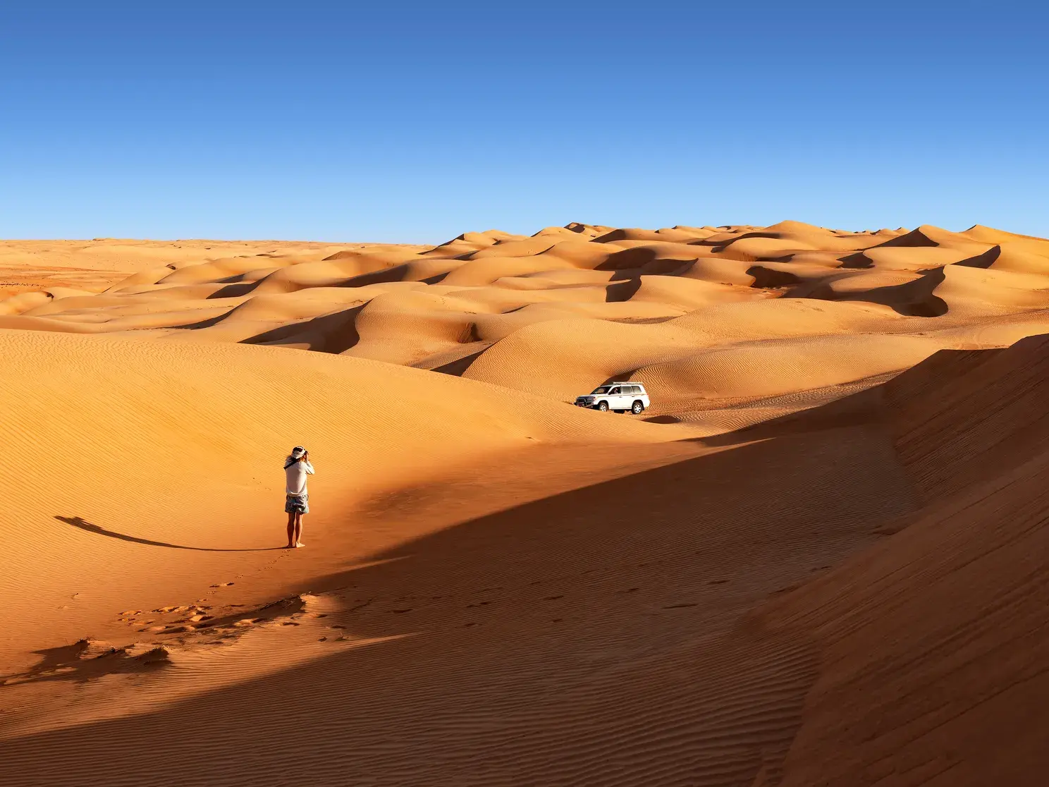 Ein Gast einer Kleingruppenreise fotografiert die goldenen Dünen der Wahiba Sands Wüste im Oman, im Hintergrund ein Geländewagen.