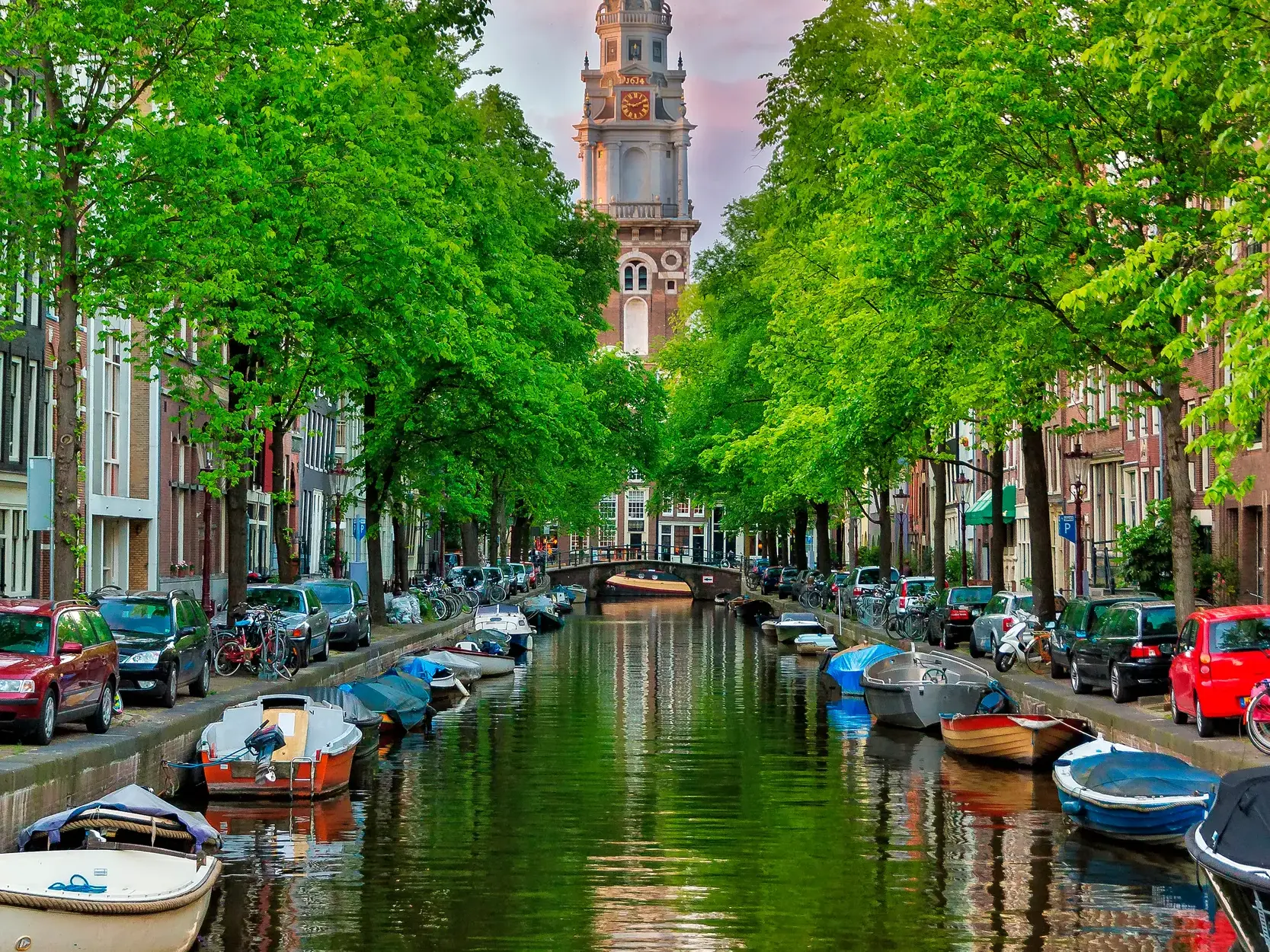 Idyllische Gracht im Amsterdamer Grachtengürtel mit am Ufer liegenden Booten, umsäumt von grünen Bäumen und historischen Häuserfassaden.