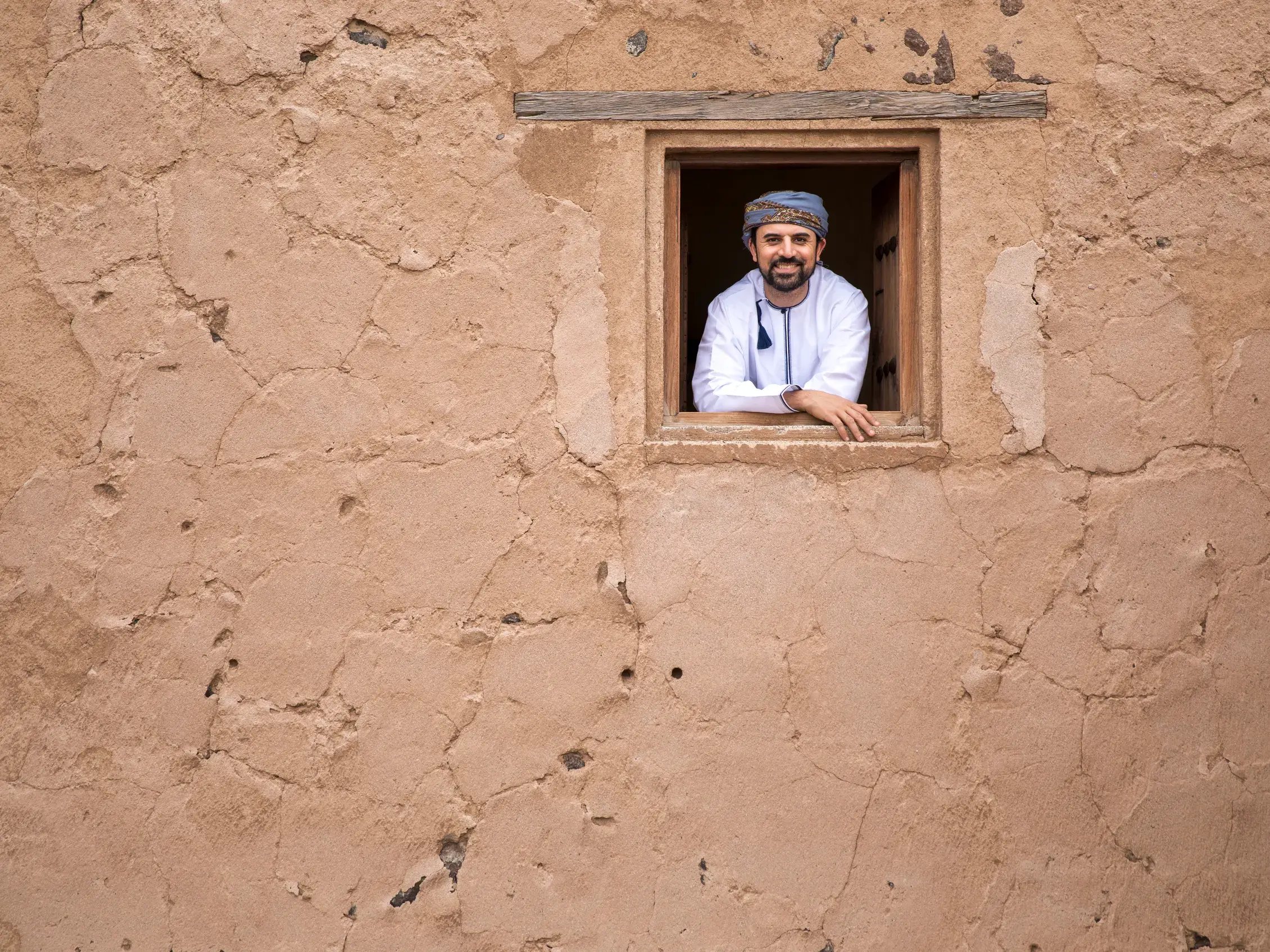 Ein lächelnder Omani in traditioneller Kleidung lehnt in einem Fenster einer Lehmfestung in der Bergoase Al-Hamra.
