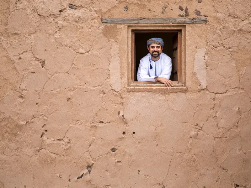 Ein lächelnder Omani in traditioneller Kleidung lehnt in einem Fenster einer Lehmfestung in der Bergoase Al-Hamra.