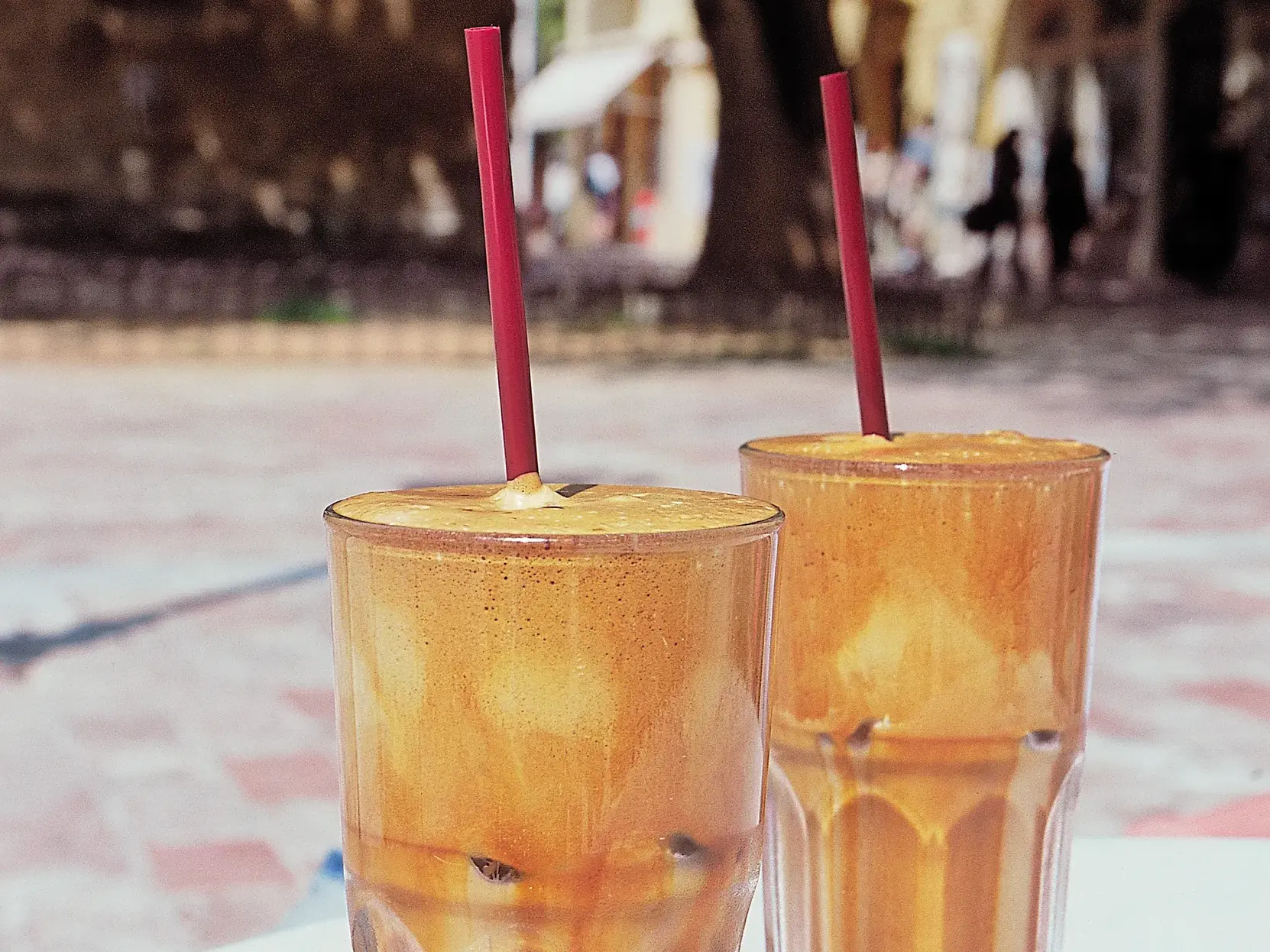 Zwei hohe Gläser mit eisgekühltem Frappé und roten Trinkhalmen stehen auf einem Tisch in einer Taverne auf Korfu.