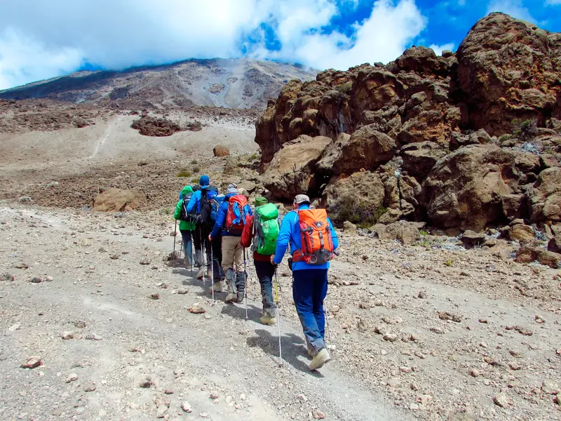 Eine kleine Trekkinggruppe wandert mit Stöcken auf einem steinigen Pfad durch die karge Vulkanlandschaft des Kilimanjaro.