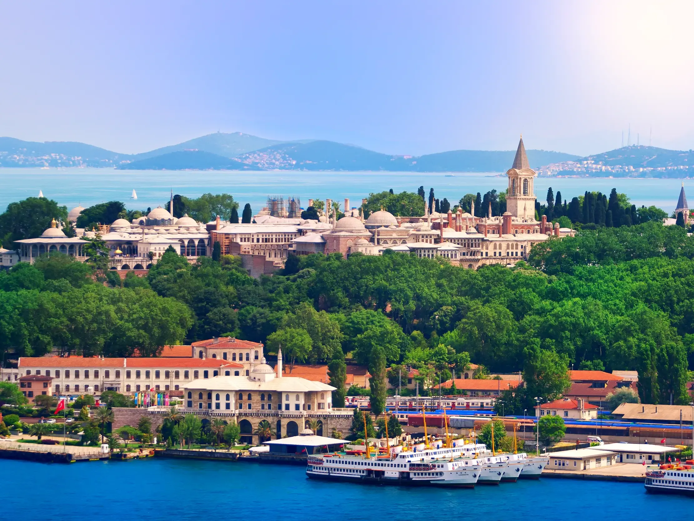 Majestätischer Blick vom Bosporus auf den Topkapi-Palast in Istanbul, umgeben von grünen Gärten am Ufer bei Sonnenschein.