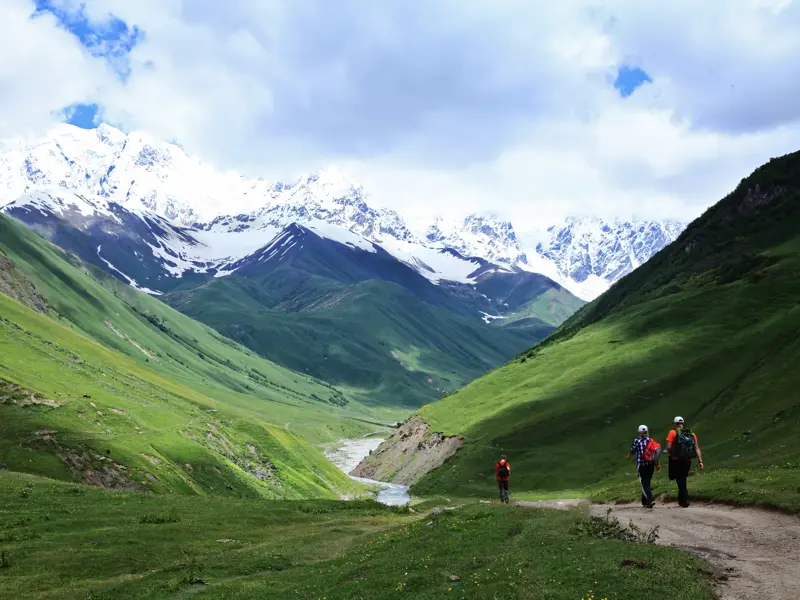 Eine kleine Wandergruppe beim Trekking in einem grünen Tal in Georgien mit Blick auf die schneebedeckten Gipfel des Kaukasus.