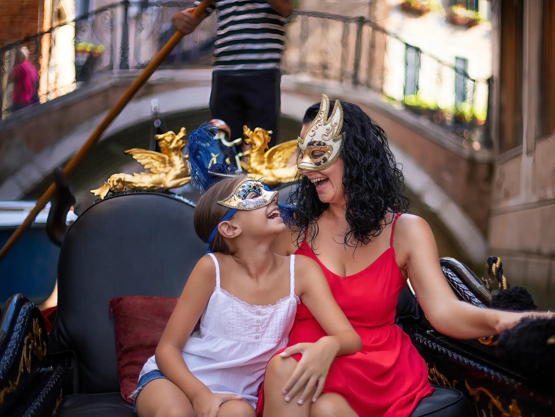 Mutter und Tochter mit venezianischen Masken genießen eine fröhliche Gondelfahrt durch einen engen Kanal in Venedig.
