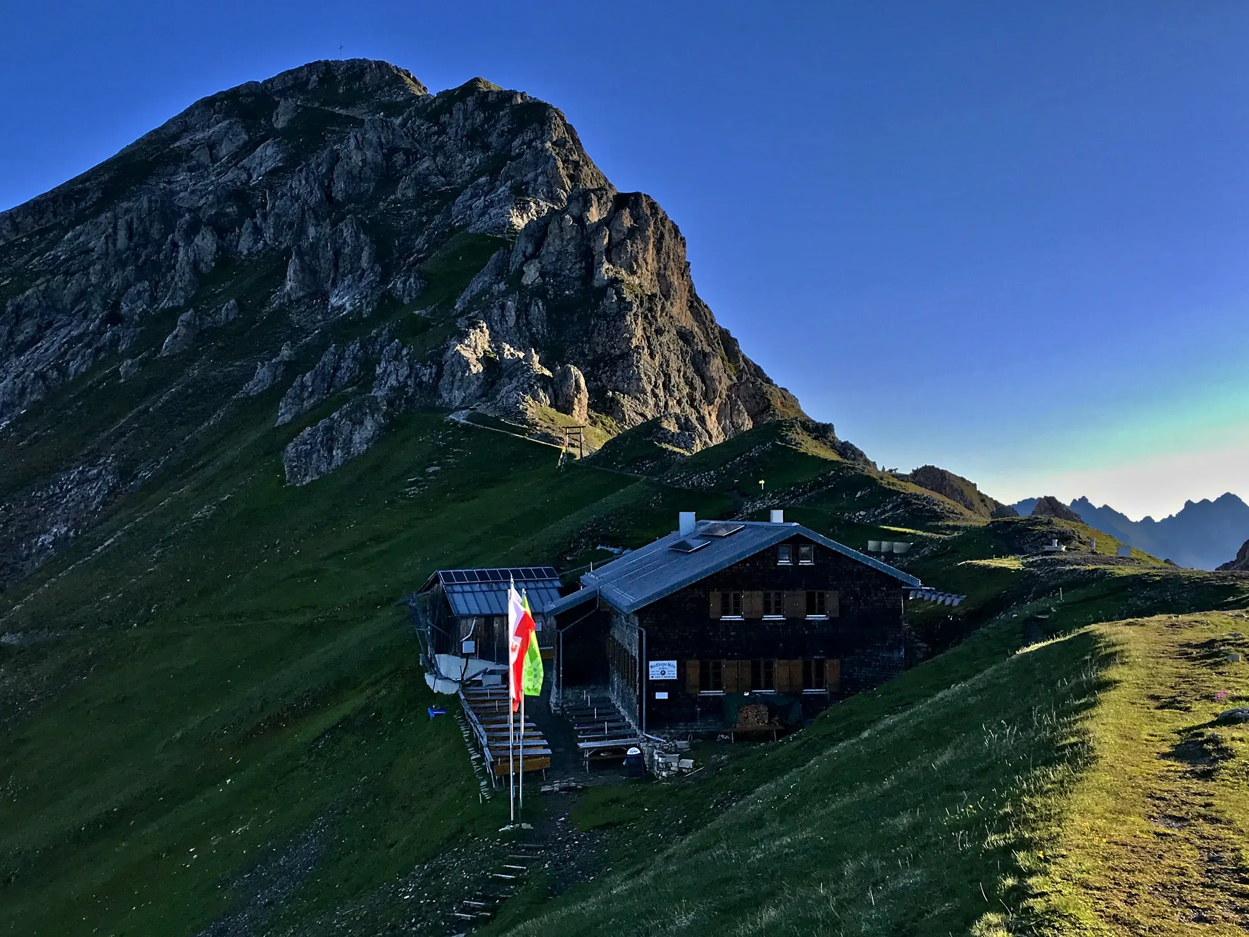 Die rustikale Nördlinger Hütte im Karwendelgebirge, an einen grünen Berghang geschmiegt unter der felsigen Reither Spitze.