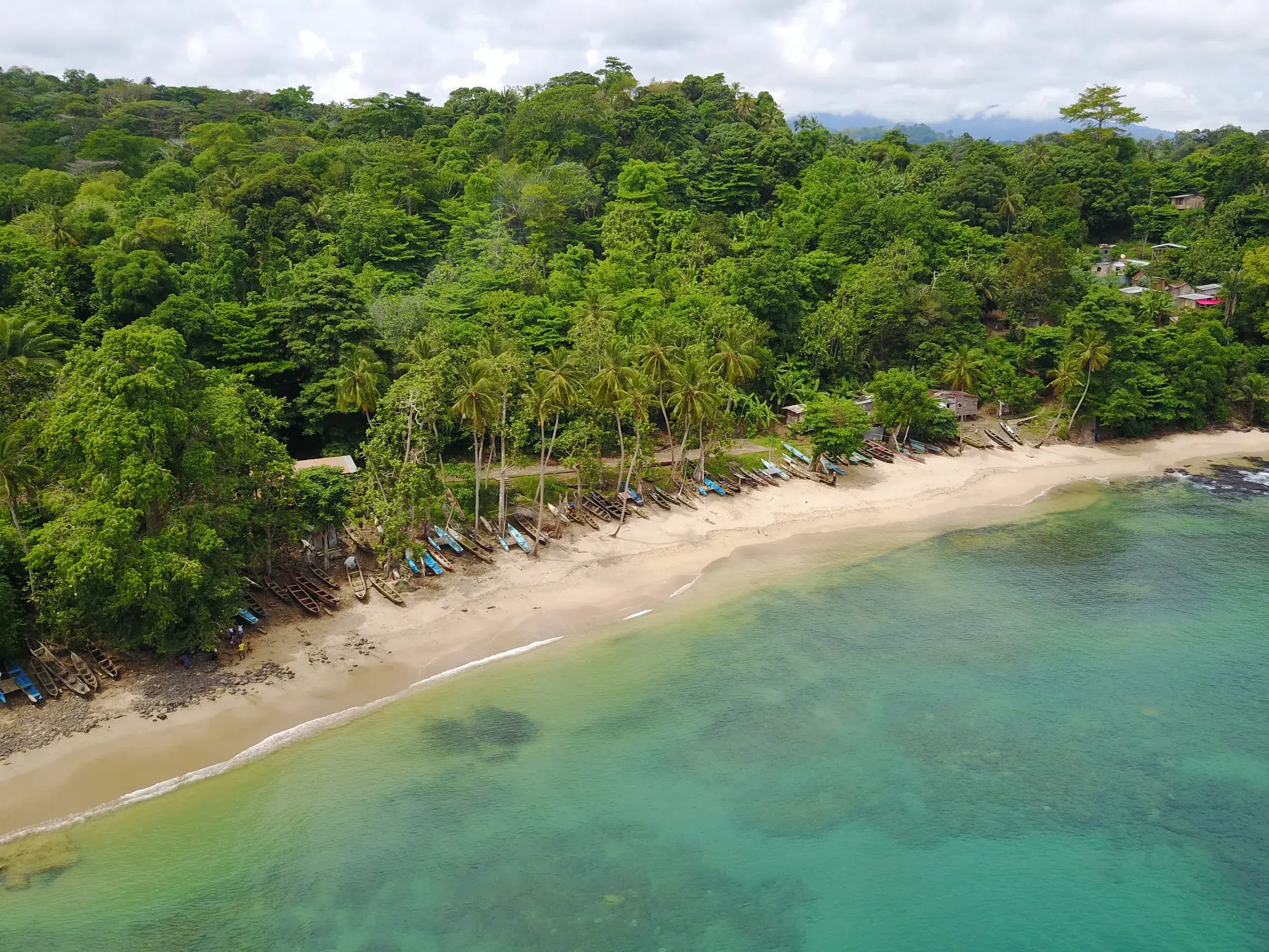Bunte Fischerboote liegen an einem abgelegenen Sandstrand auf São Tomé, eingerahmt von üppigem Regenwald und dem Atlantik.