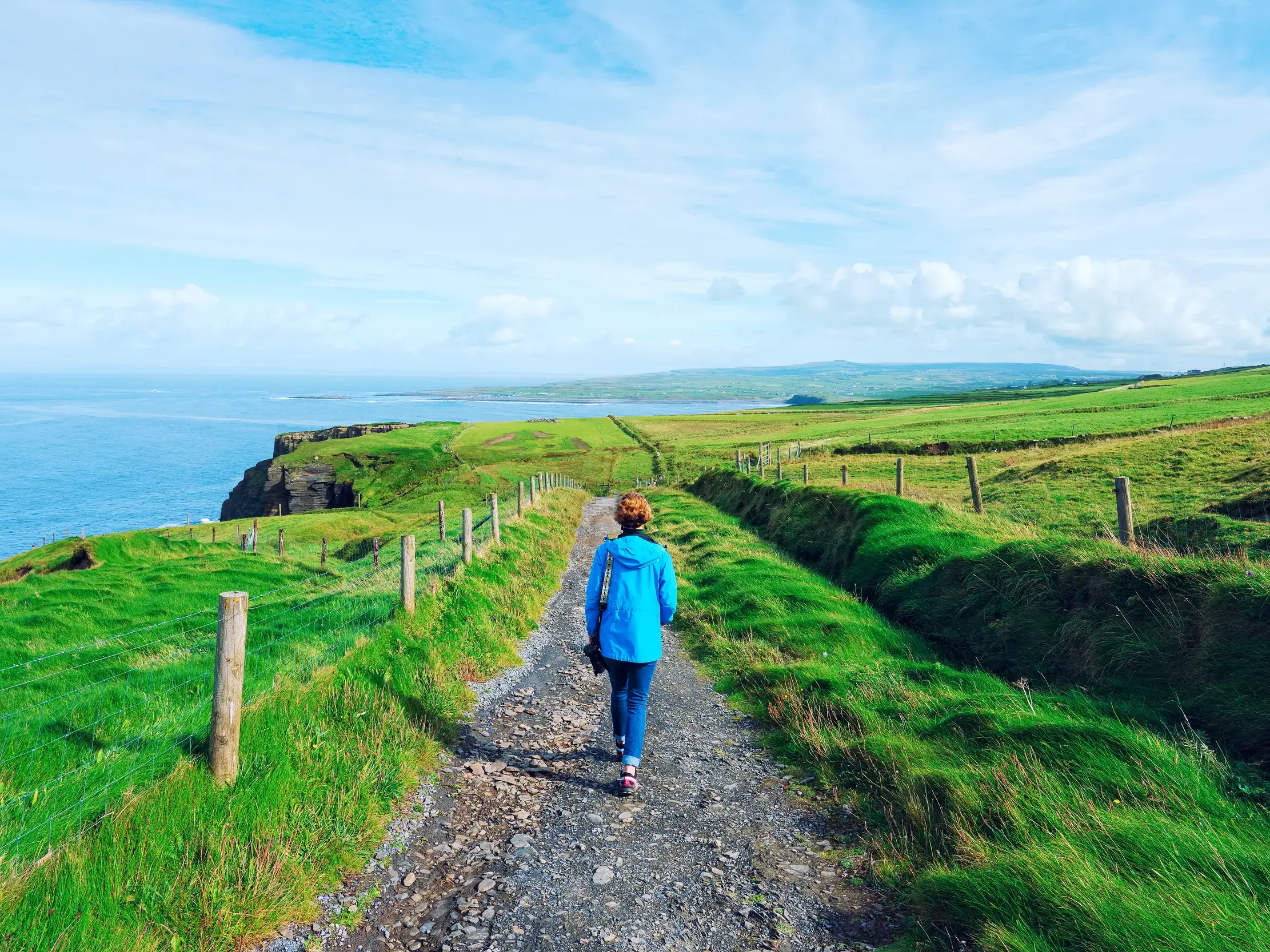 Eine Frau in blauer Jacke wandert auf einem Küstenpfad entlang der beeindruckenden, grünen Klippen von Moher in Irland.