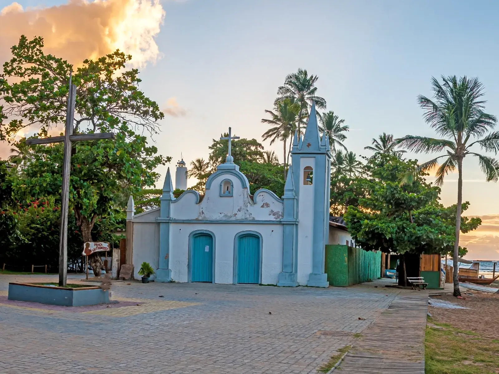 Die weiße Kirche São Francisco de Assis mit blauen Türen am Strand von Praia do Forte umgeben von Palmen bei Sonnenuntergang.