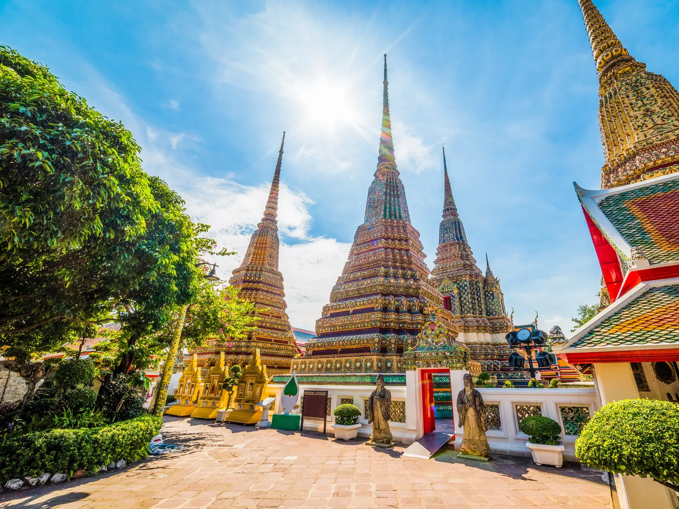 Imposante, goldverzierte Chedis der Tempelanlage Wat Pho in Bangkok ragen unter strahlender Sonne in den blauen Himmel.