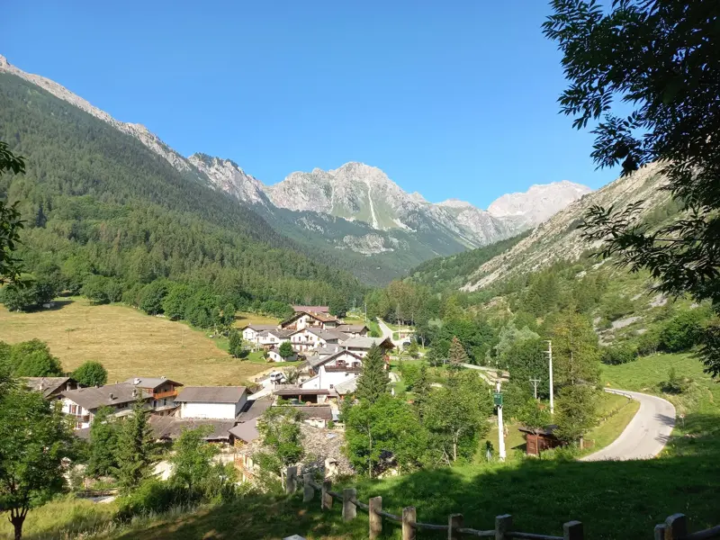 Panoramablick auf ein abgeschiedenes Bergdorf im sonnigen Maira-Tal, umgeben von grünen Hängen und felsigen Alpengipfeln.
