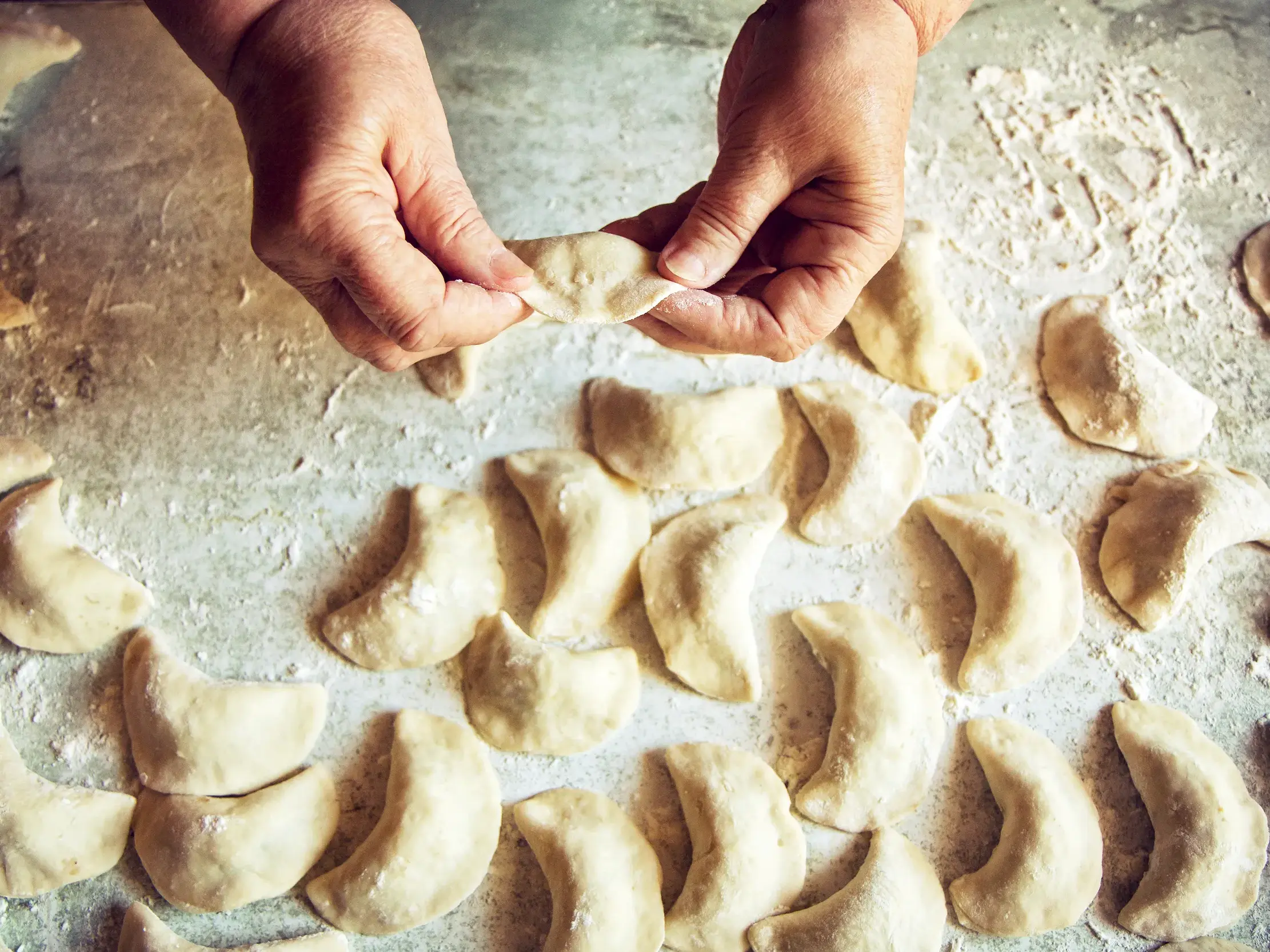 Ältere Hände formen sorgfältig traditionelle polnische Piroggen, gefüllte Teigtaschen, auf einer bemehlten Arbeitsfläche.