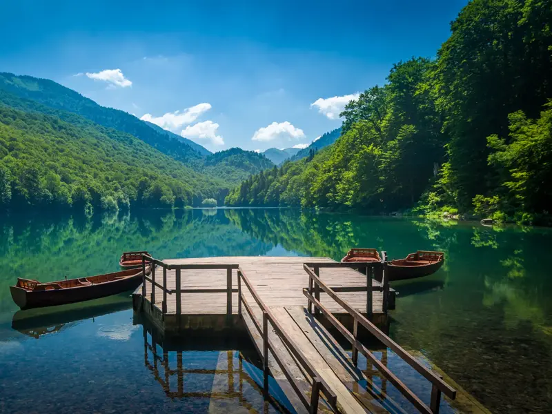Ein Holzsteg mit Ruderbooten am spiegelglatten Biogradsko See, umgeben von den dichten Wäldern des Nationalparks in Montenegro.