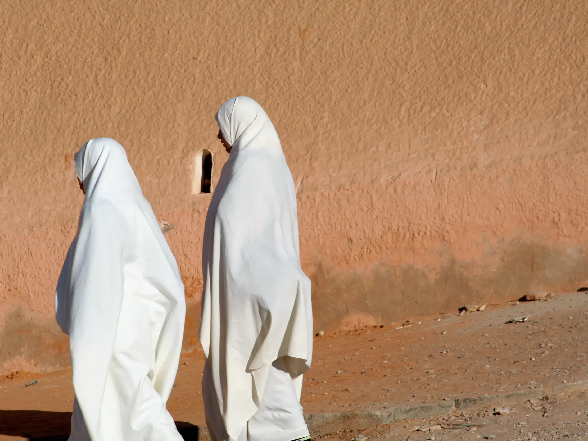 Zwei Frauen in traditionellen weißen Haiks gehen an einer ockerfarbenen Lehmwand in der Oasenstadt Ghardaia in Algerien entlang.