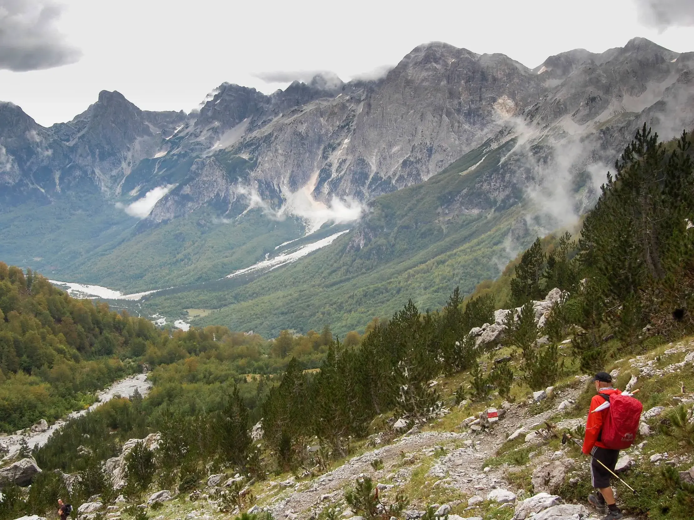 Wanderer mit Rucksack auf dem steinigen Peaks of the Balkans Trail mit Aussicht auf das Valbonatal und die Albanischen Alpen.