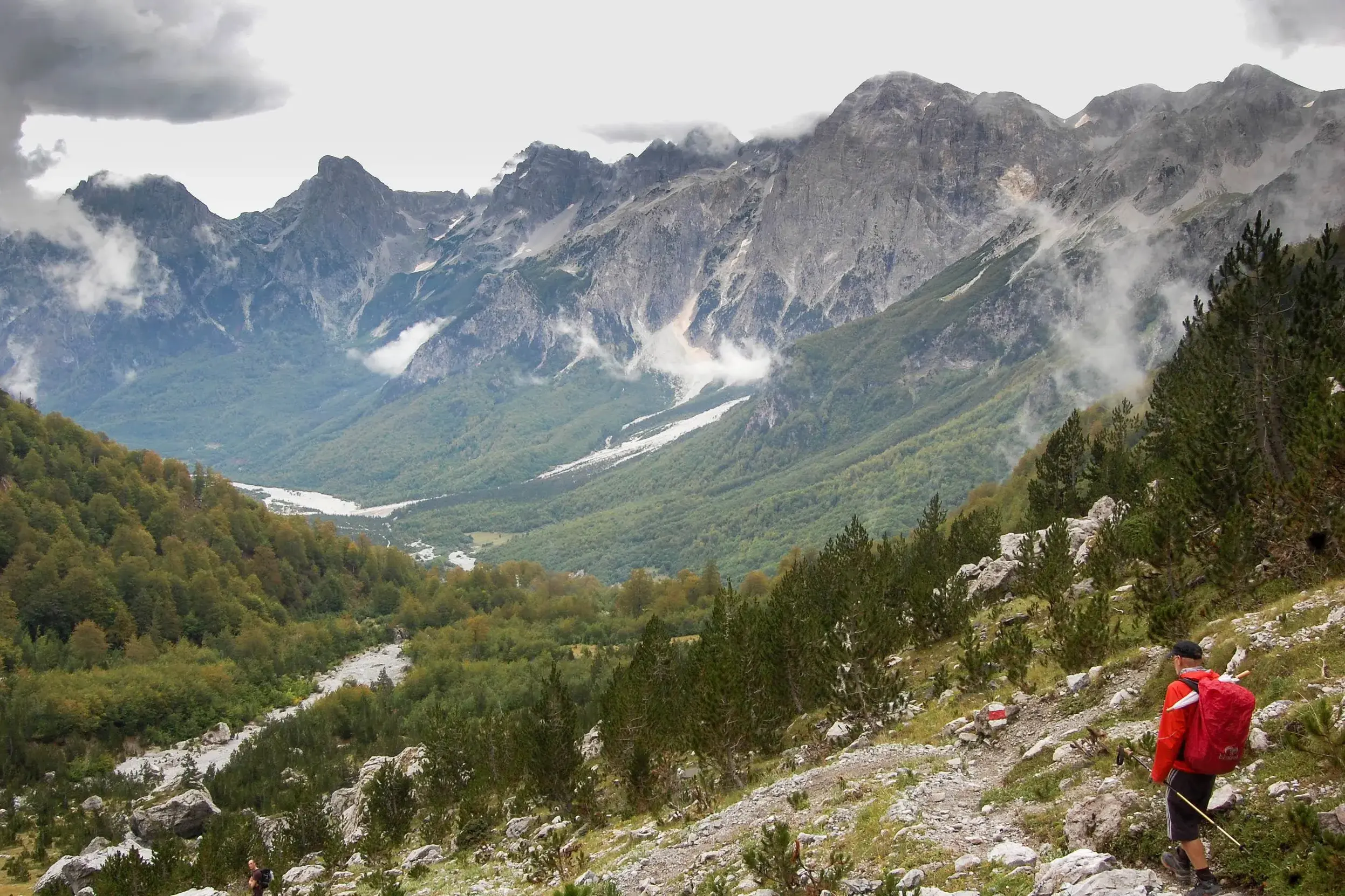 Ein Wanderer mit rotem Rucksack beim Trekking auf einem Bergpfad mit Panoramablick über das grüne Valbonatal in Albanien.