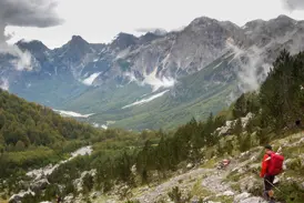 Ein Wanderer mit rotem Rucksack beim Trekking auf einem Bergpfad mit Panoramablick über das grüne Valbonatal in Albanien.