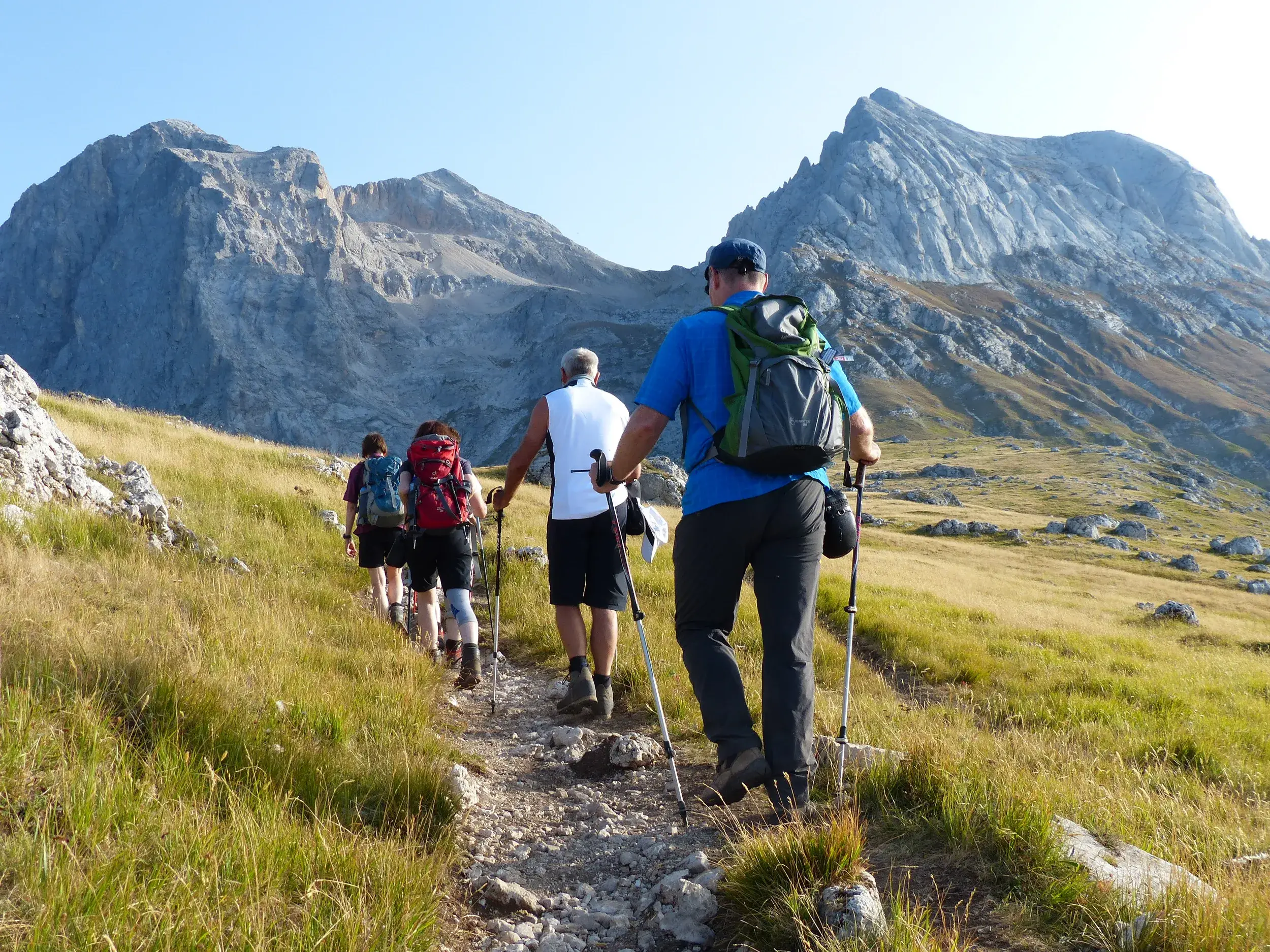 Reisegruppe beim Trekking auf einem schmalen Pfad durch eine grüne Hochebene mit Blick auf die Gipfel des Gran Sasso Massivs.