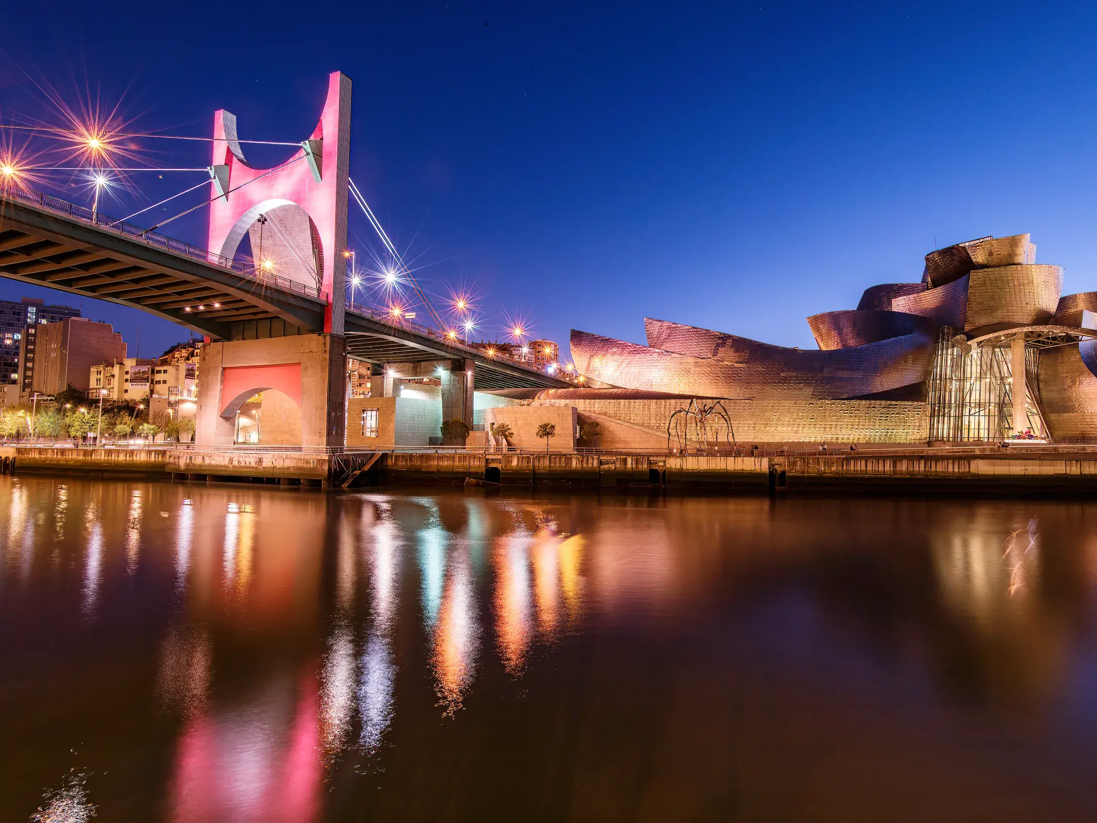 Moderne Architektur in Bilbao: Das Guggenheim-Museum am Rio Nervión bei Nacht mit beleuchteter Brücke und Spiegelung im Wasser.