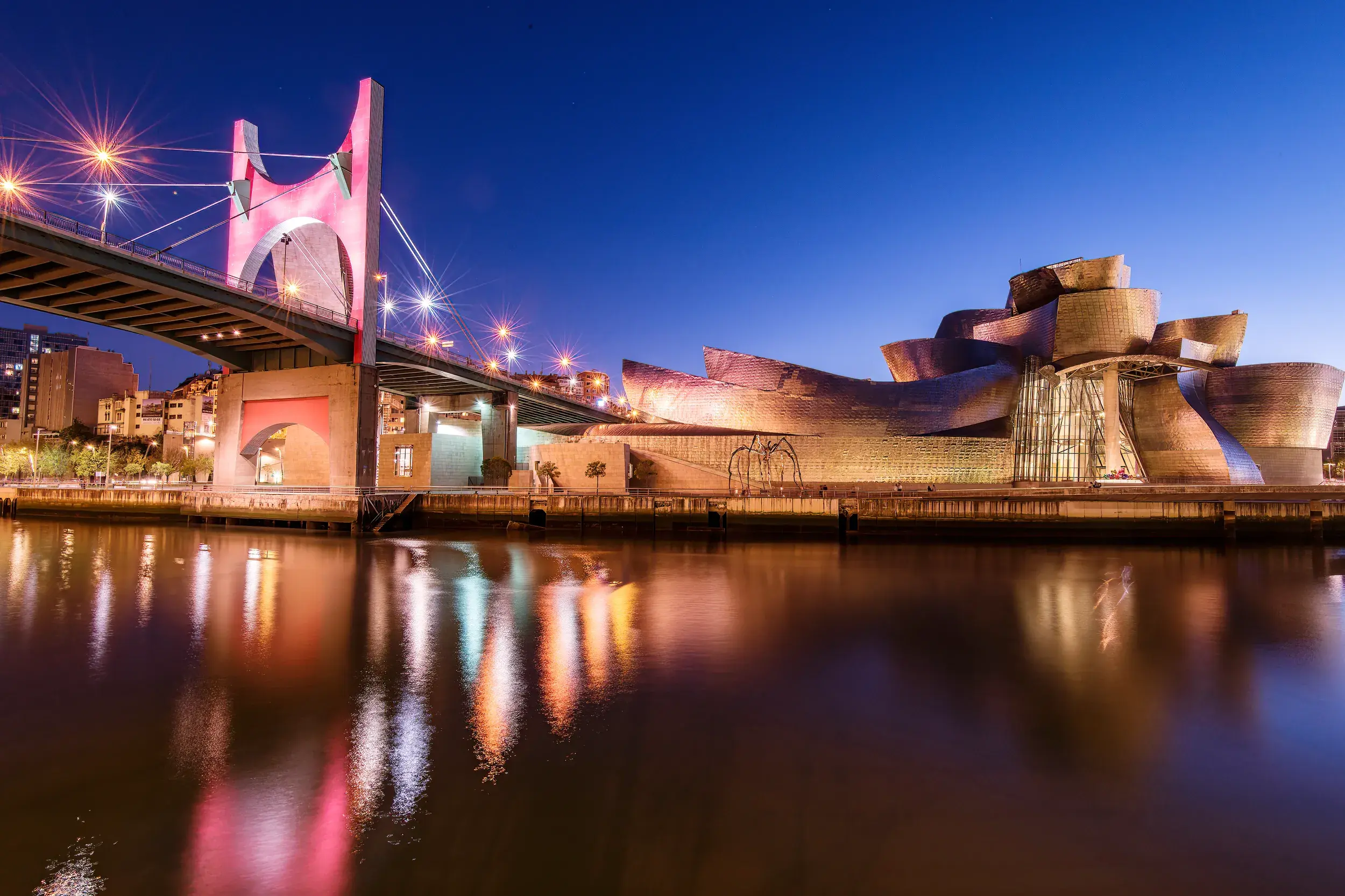 Moderne Architektur in Bilbao: Das Guggenheim-Museum am Rio Nervión bei Nacht mit beleuchteter Brücke und Spiegelung im Wasser.