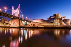 Moderne Architektur in Bilbao: Das Guggenheim-Museum am Rio Nervión bei Nacht mit beleuchteter Brücke und Spiegelung im Wasser.