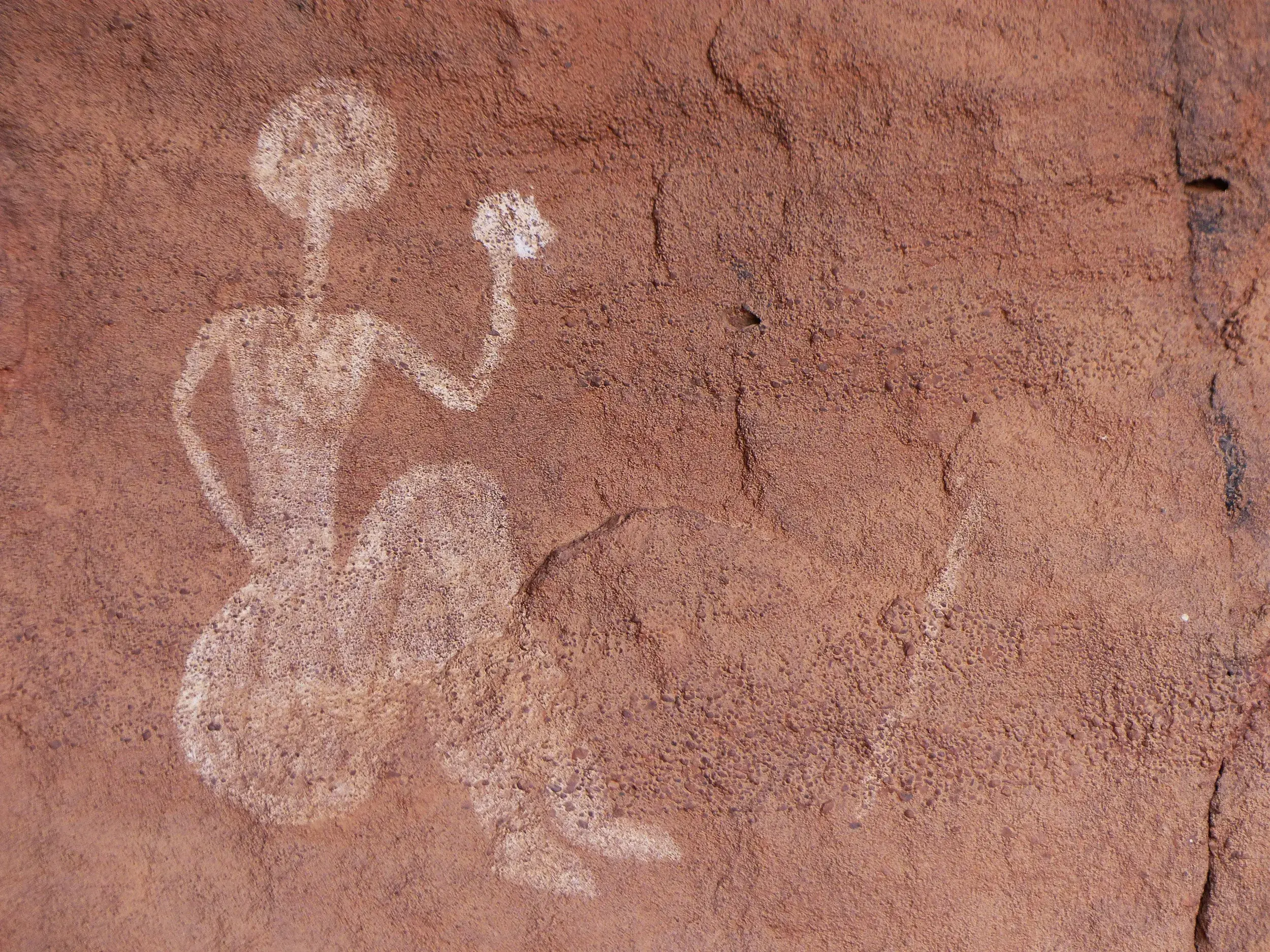 Prähistorische Felsmalerei im Tassili n'Ajjer Nationalpark zeigt eine weiße, stilisierte Figur an einer roten Felswand.