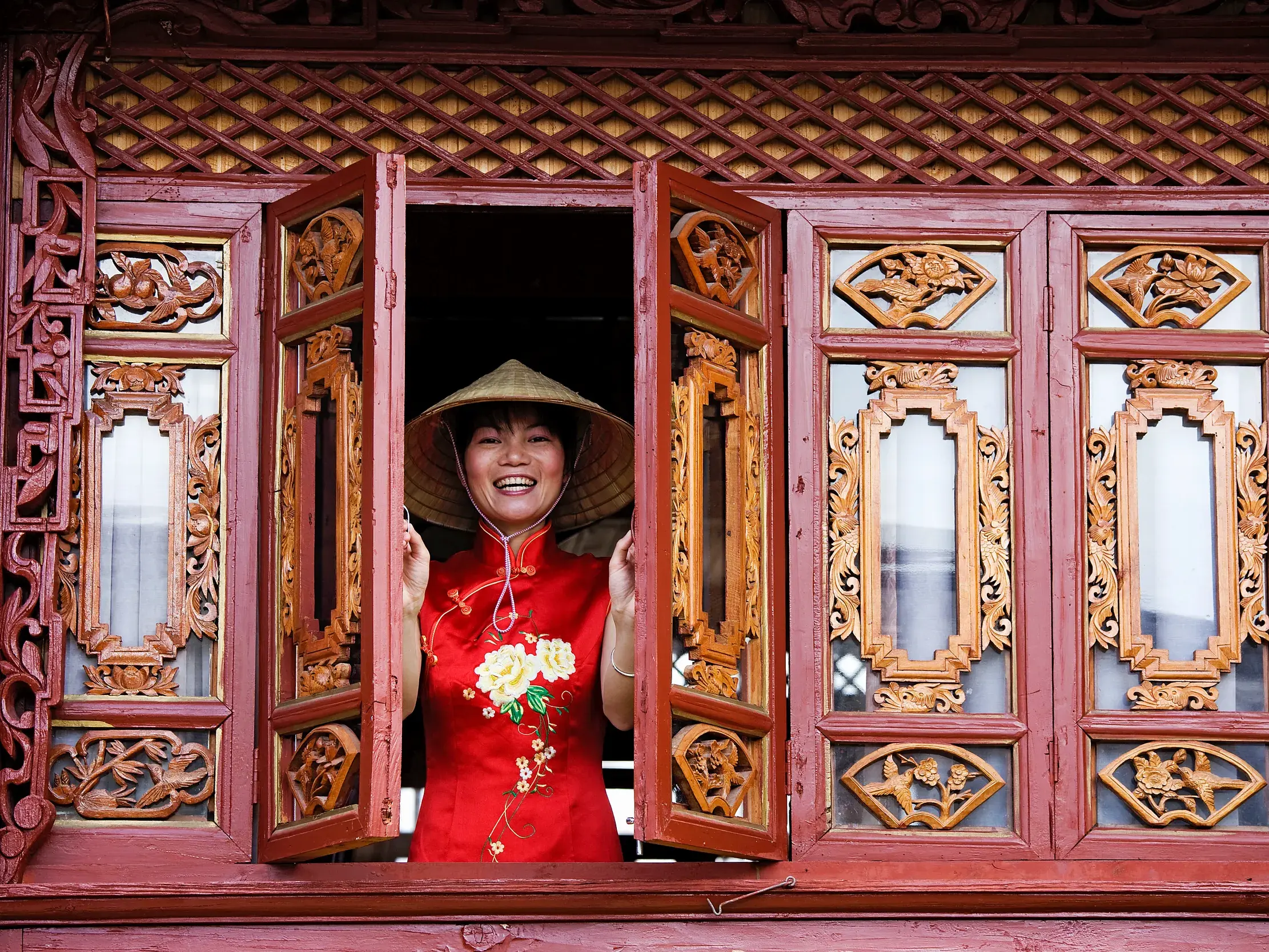 Eine Frau in roter traditioneller Kleidung und Hut blickt lächelnd aus dem reich verzierten Fenster eines alten Hauses in Lijiang.