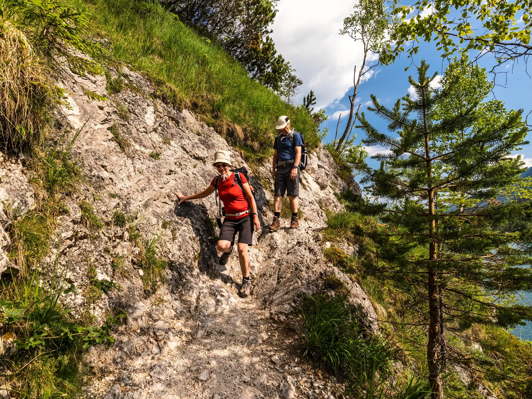Zwei Wanderer auf einem schmalen, felsigen Bergpfad an einem sonnigen Berghang in den bayerischen Voralpen.