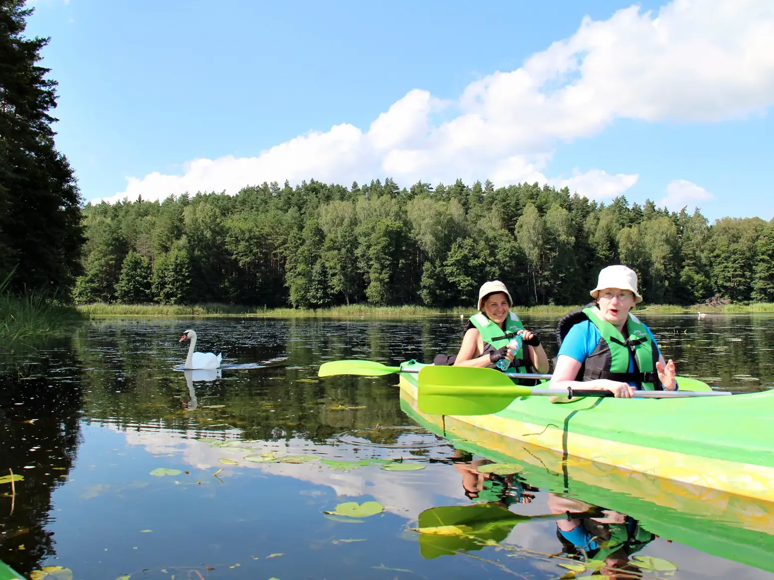 Zwei Frauen in einem Kajak bei einer sonnigen Tour auf dem Fluss Krutynia in Masuren, vorbei an einem Schwan und dichten Wäldern.