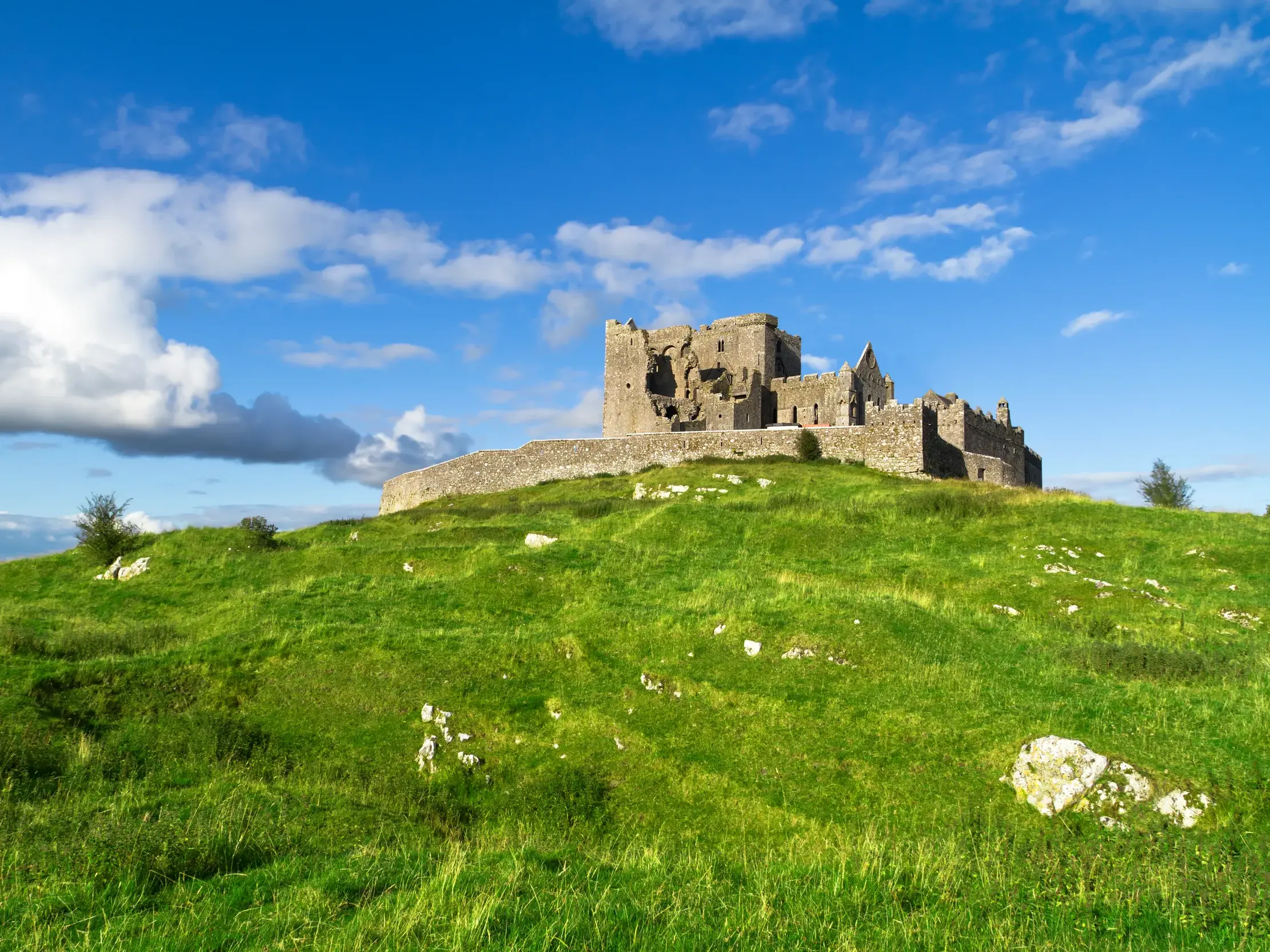Die felsgrauen Ruinen des Burgbergs von Cashel thronen auf einem grünen Hügel in Irland unter blauem Himmel mit Wolken.