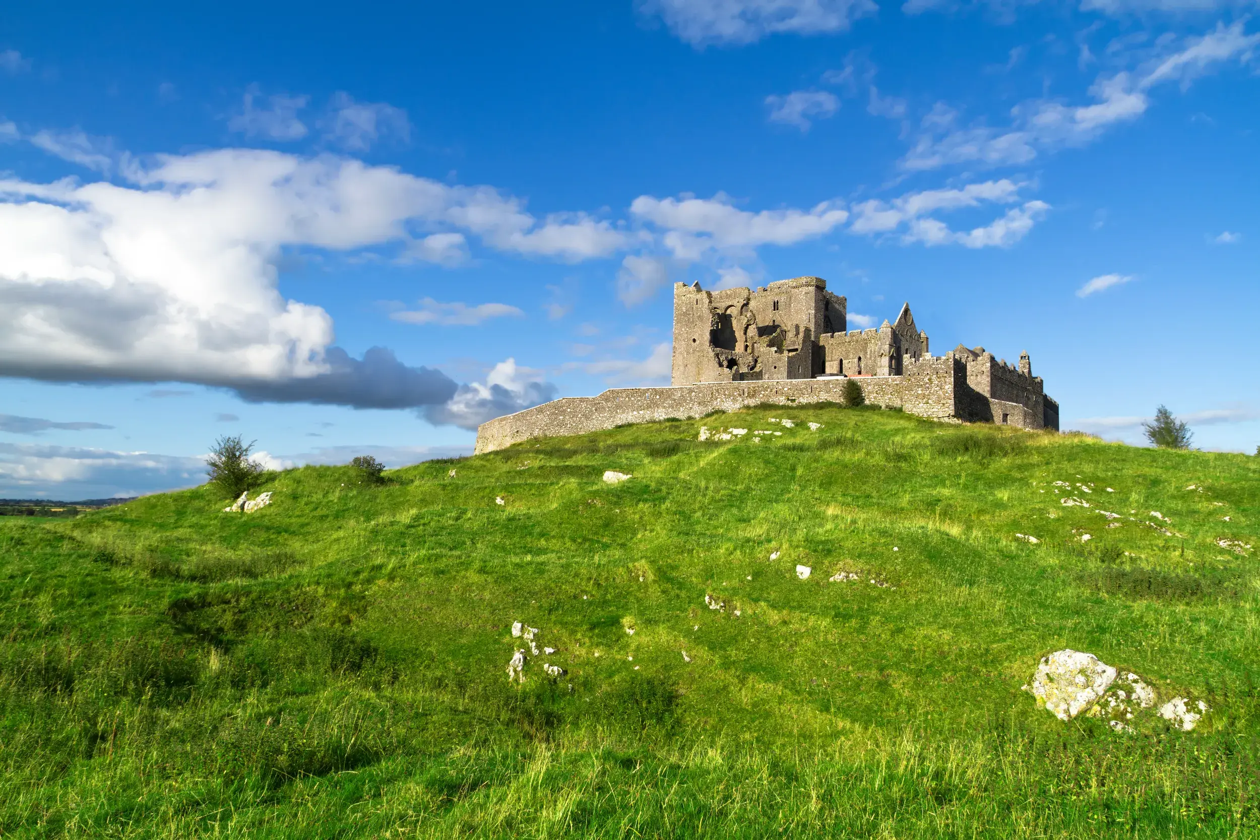 Die felsgrauen Ruinen des Burgbergs von Cashel thronen auf einem grünen Hügel in Irland unter blauem Himmel mit Wolken.