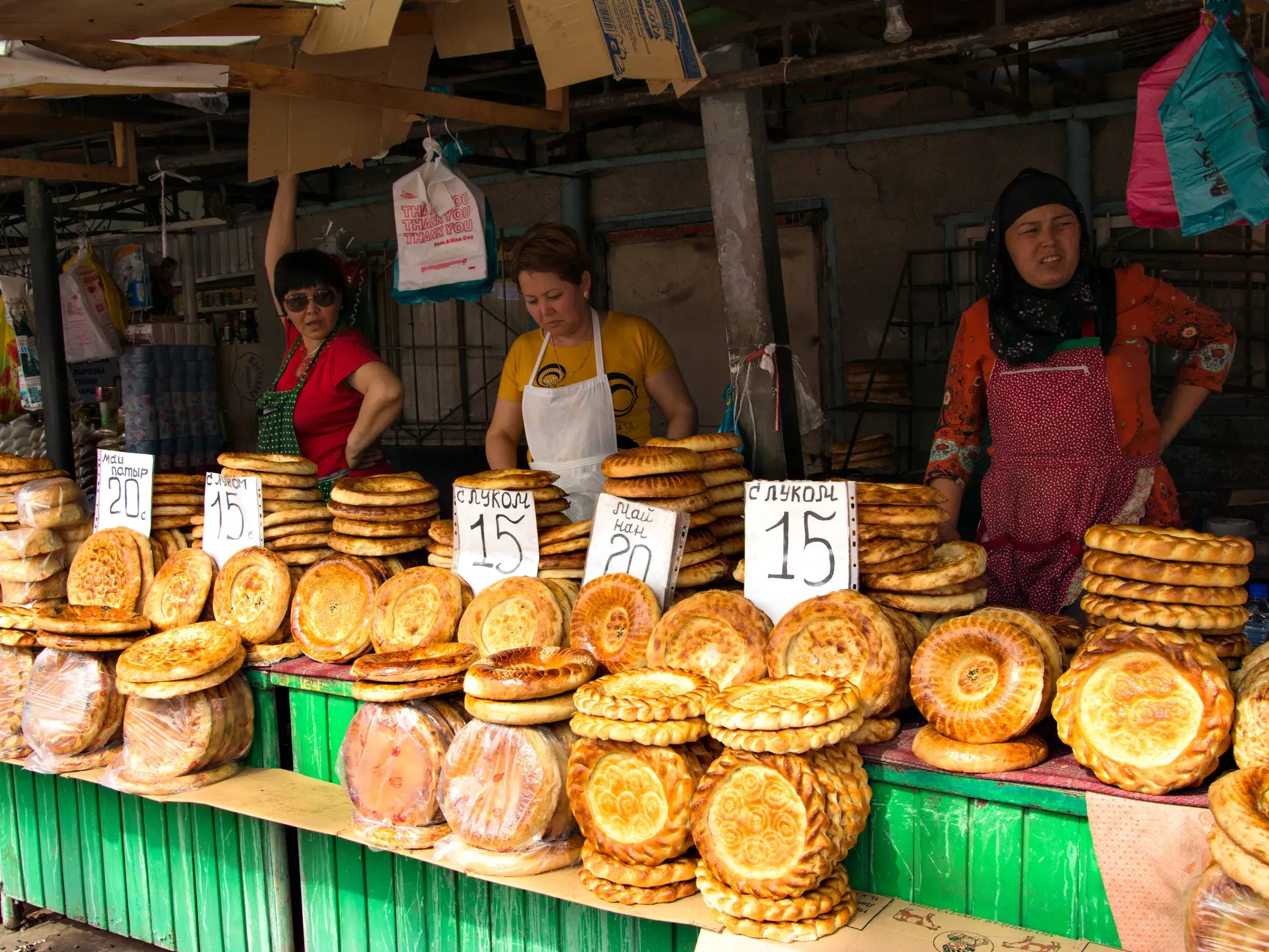 Drei kirgisische Frauen verkaufen traditionelles, verziertes Fladenbrot an einem bunten Marktstand auf dem Osch-Basar in Bischkek.