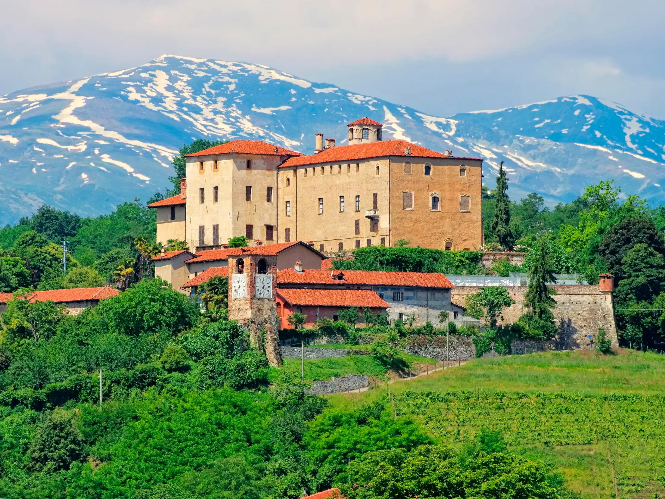 Das Castello della Manta thront auf einem grünen Hügel im Piemont, im Hintergrund der schneebedeckte Gipfel des Monviso.