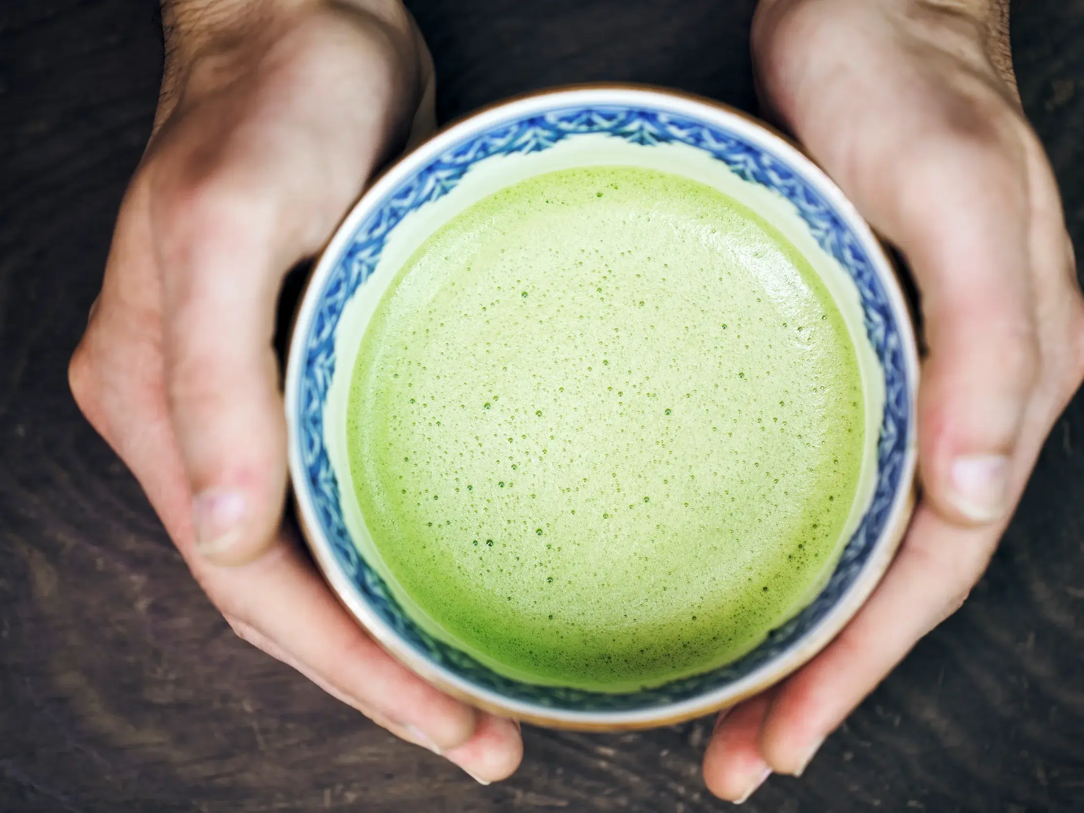 Zwei Hände halten eine traditionelle Schale mit schaumigem, grünem Matcha-Tee bei einer Teeprobe in Japan.