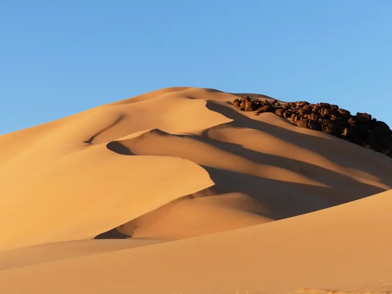 Ein scharf gezeichneter Dünenkamm im Erg Admer, dessen goldener Sand an dunkle Felsen der Sahara grenzt.