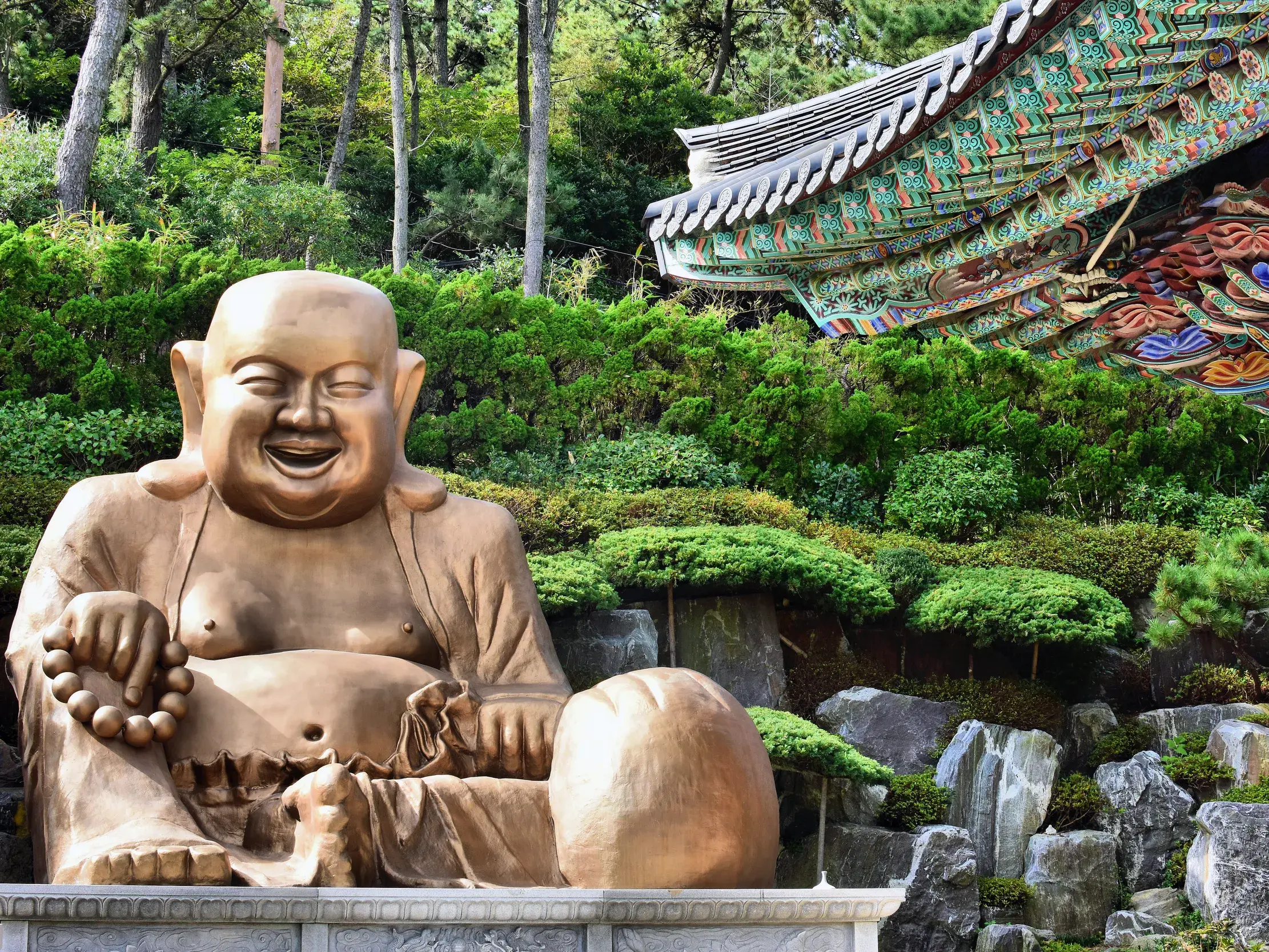 Große, lachende Buddha-Statue in einem Tempelgarten in Busan, im Hintergrund ein traditionelles, buntes Pagodendach.